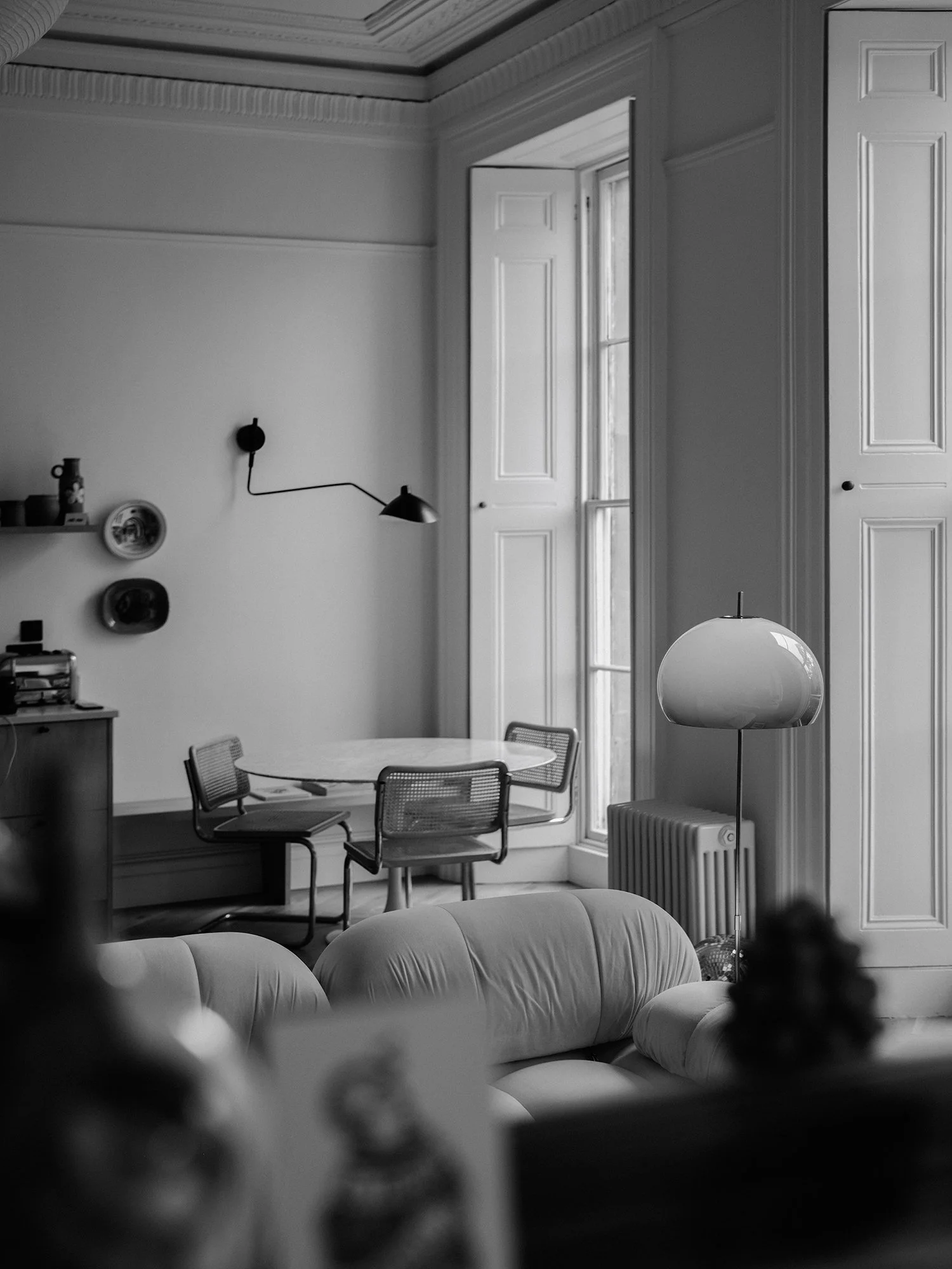 A black and white photo of a living room with a round table, chairs, a tall window with built-in shutters, a couch, a floor lamp, and a radiator.