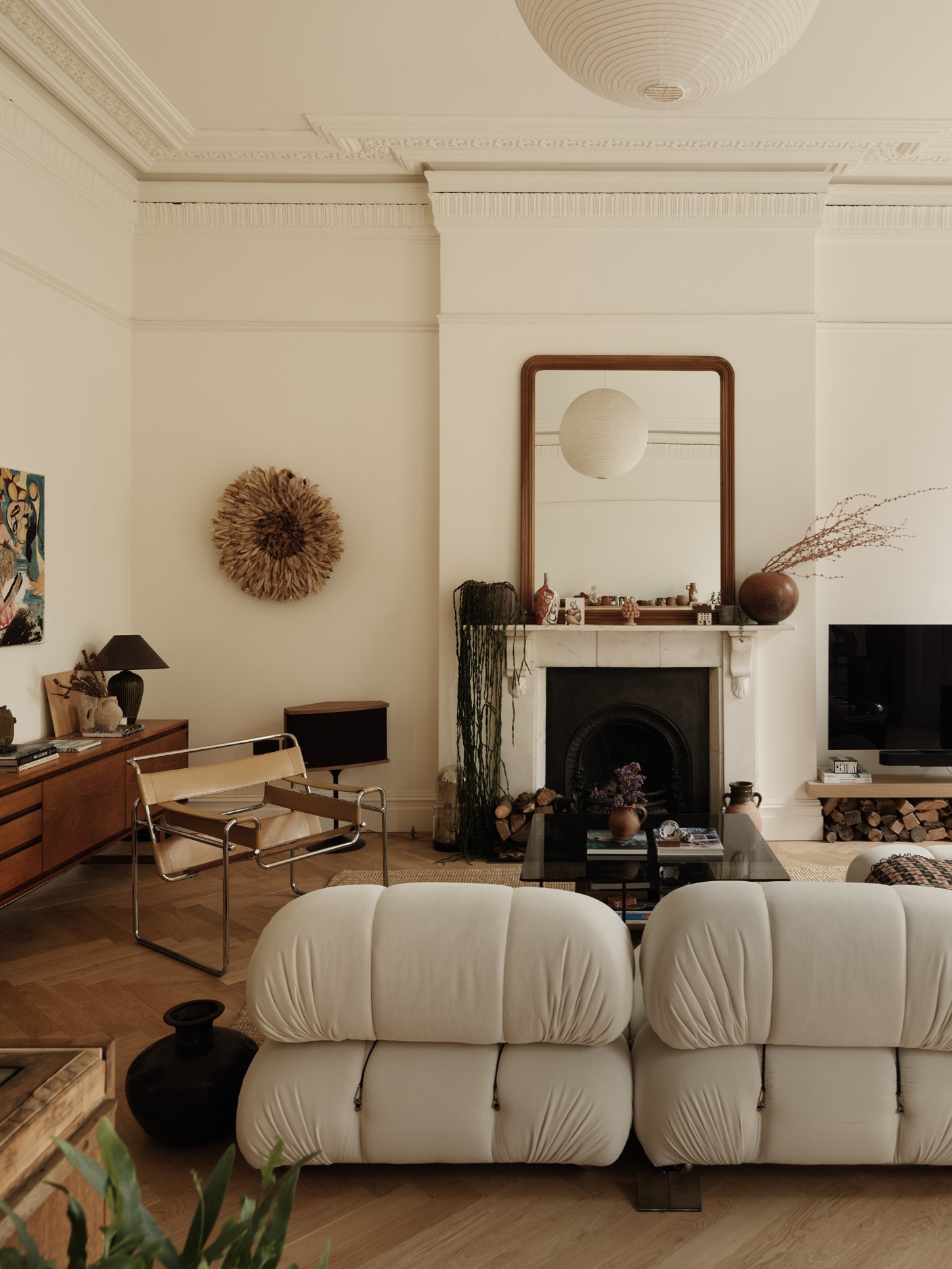 Living room with white leather sofa, fireplace with decorative items and a large mirror, wood flooring, and mid-century modern furniture. A large paper lantern hangs from the ceiling.
