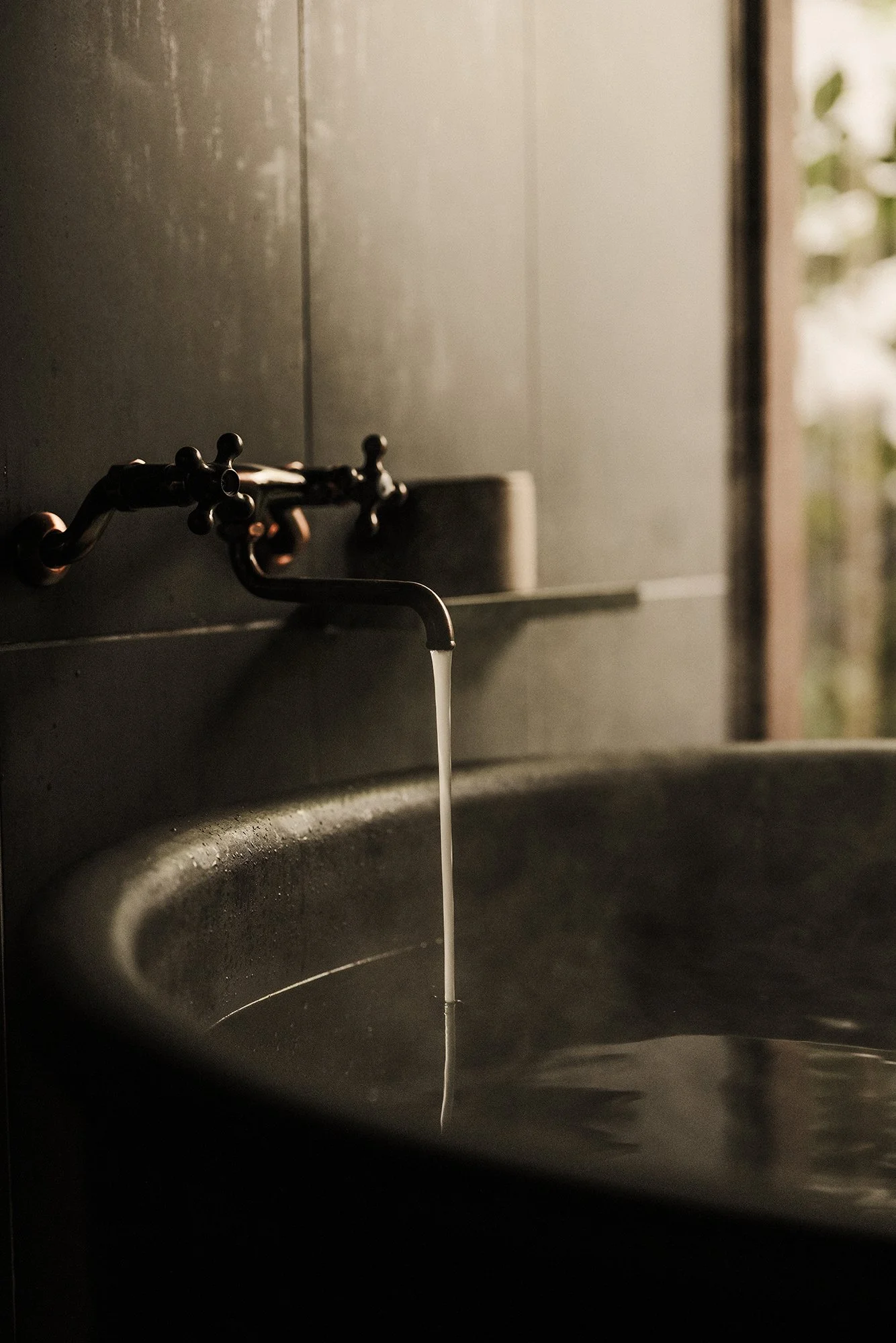 A black metal sink with a vintage-style faucet, with water flowing into it, in a dimly lit room with blurred window or door in the background.