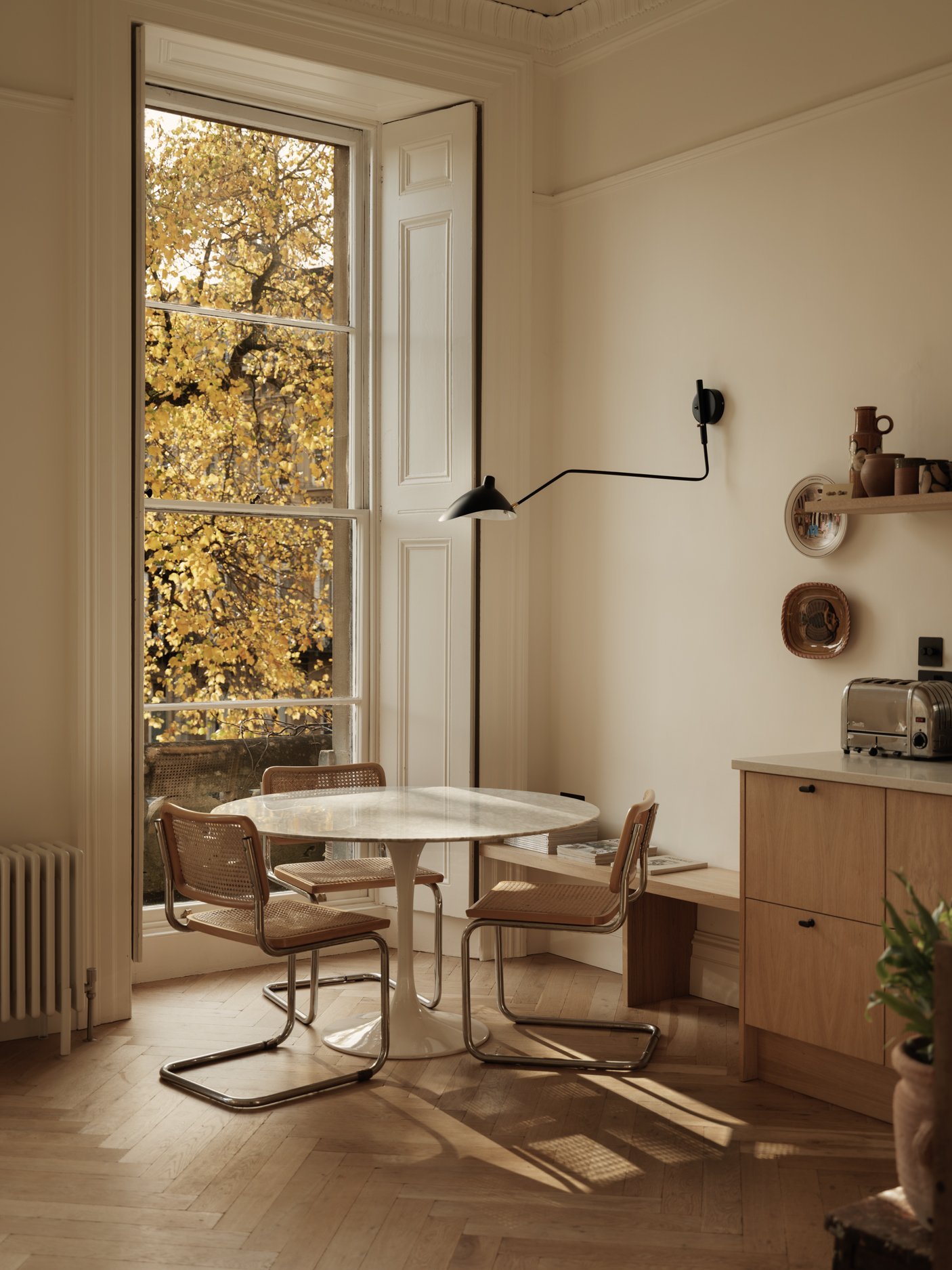 A cozy dining nook with a marble table and three wicker chairs in front of a large window with open shutters, overlooking autumn-colored trees.