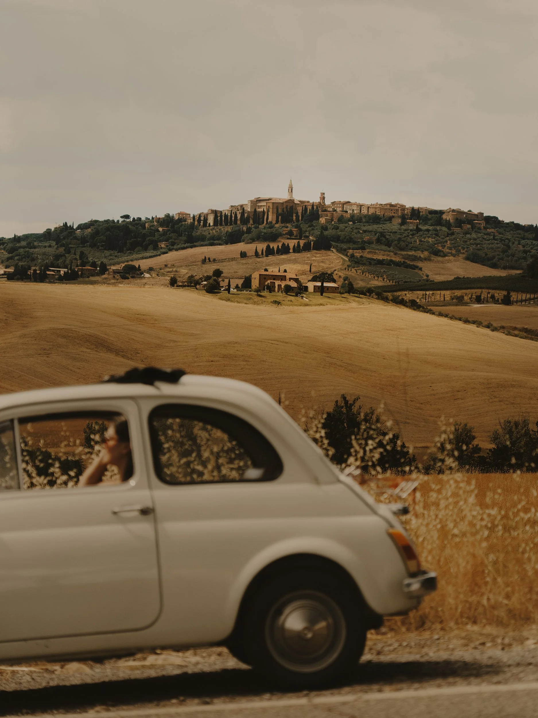 A vintage silver car driving on a rural road with a person inside, with rolling countryside and a hilltop town or village with buildings and a tower in the background.