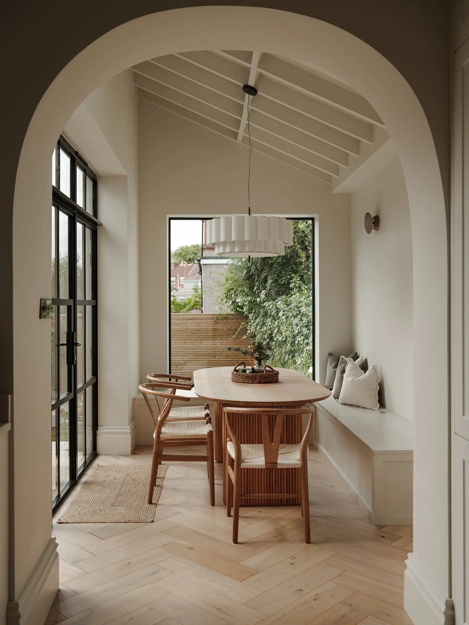 Dining area with a wooden table and four matching wooden chairs, a built-in window seat with pillows, large windows, and a modern pendant light hanging from the ceiling, with a view of outdoor greenery.