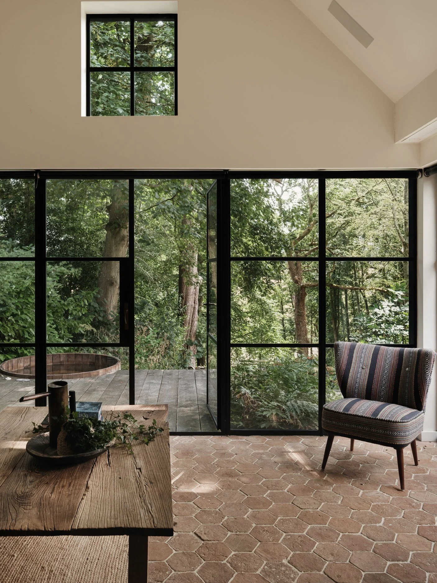 A room with a large sliding glass door looking out onto dense greenery with trees. The room has a hexagonal terracotta tiled floor, a wooden table with decorative items, and a striped upholstered chair.