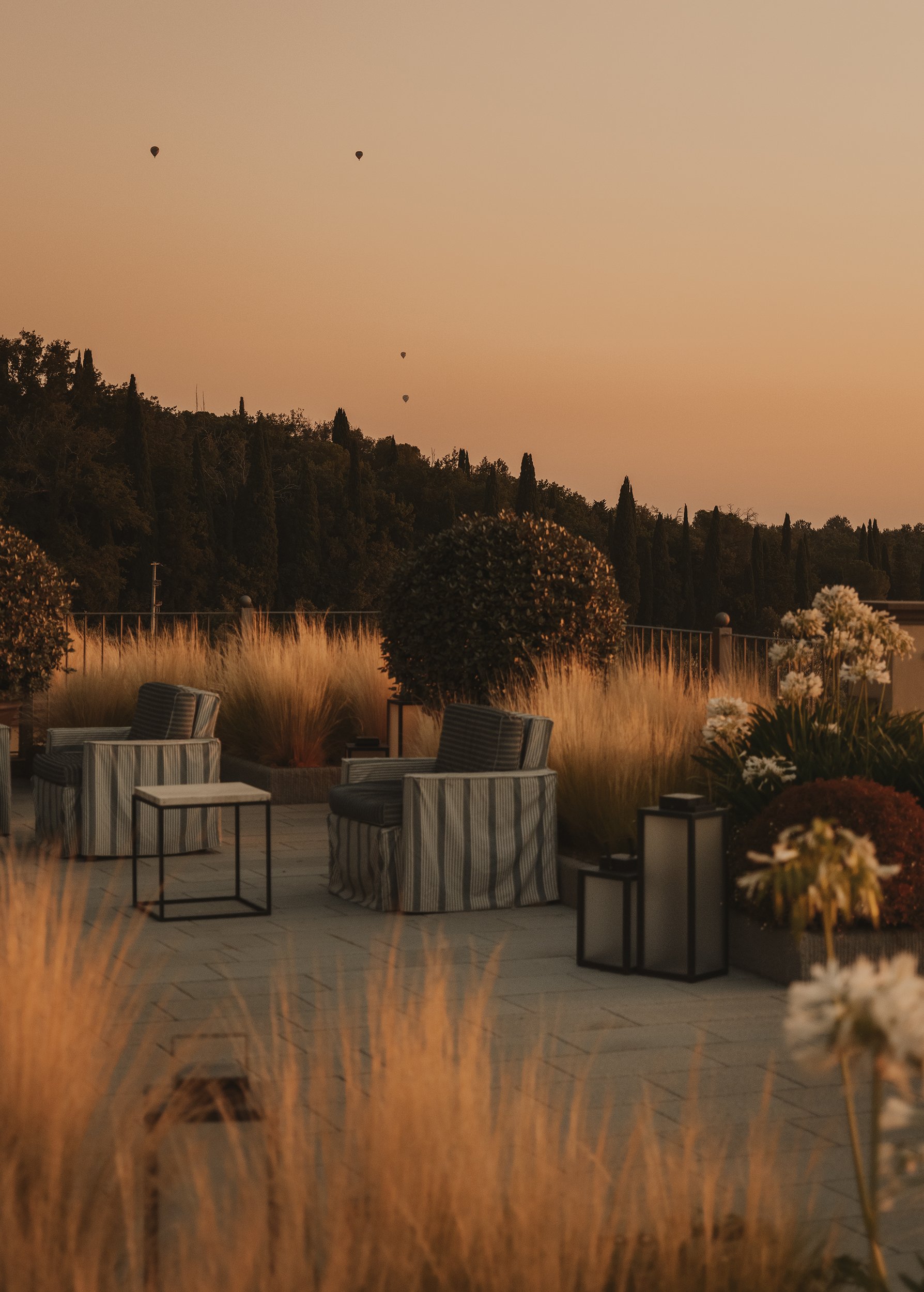 Outdoor patio seating area with striped chairs, side tables, lanterns, and decorative plants during sunset with hot air balloons in the sky.