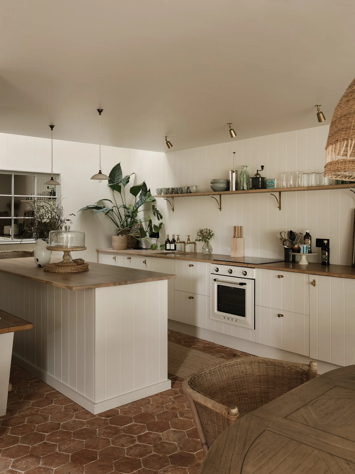 A cozy kitchen with white paneled cabinets, wooden countertops, and a terracotta tile floor. It features open shelving with cups, bowls, and glassware, as well as potted plants and various kitchen utensils.