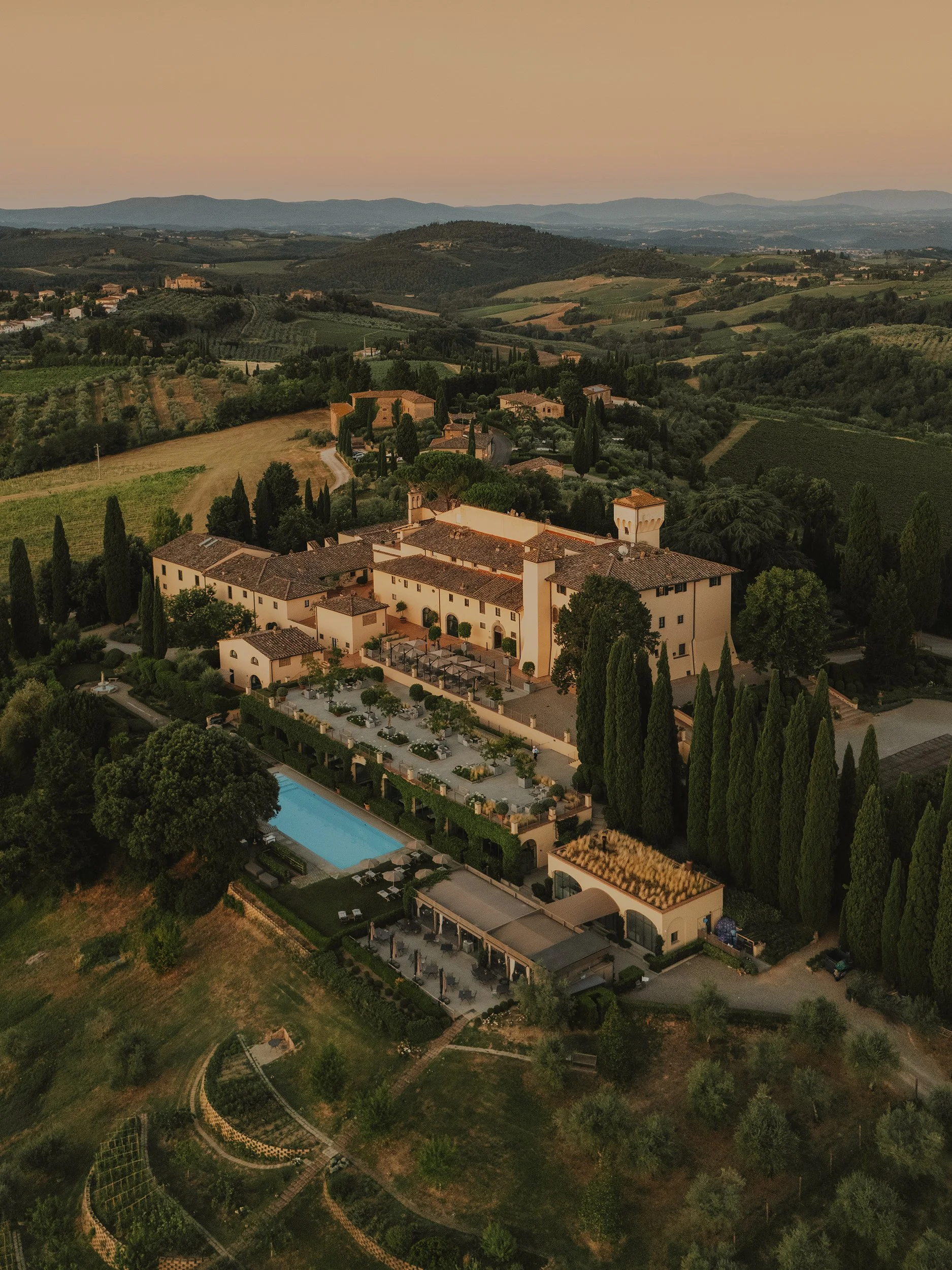 A large estate or villa in a rural landscape with rolling hills, vineyards, and trees, featuring a swimming pool and outdoor seating area.