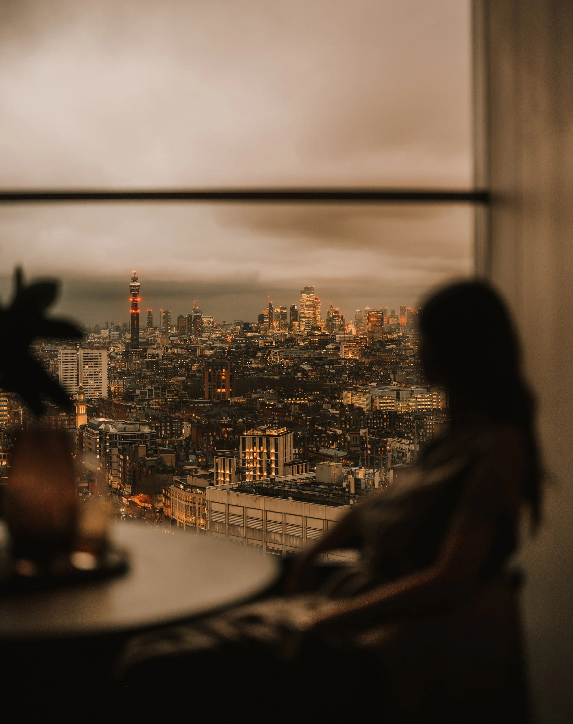 A woman sitting by a window overlooking a city skyline at dusk, with tall buildings and a tower with a red and white light.