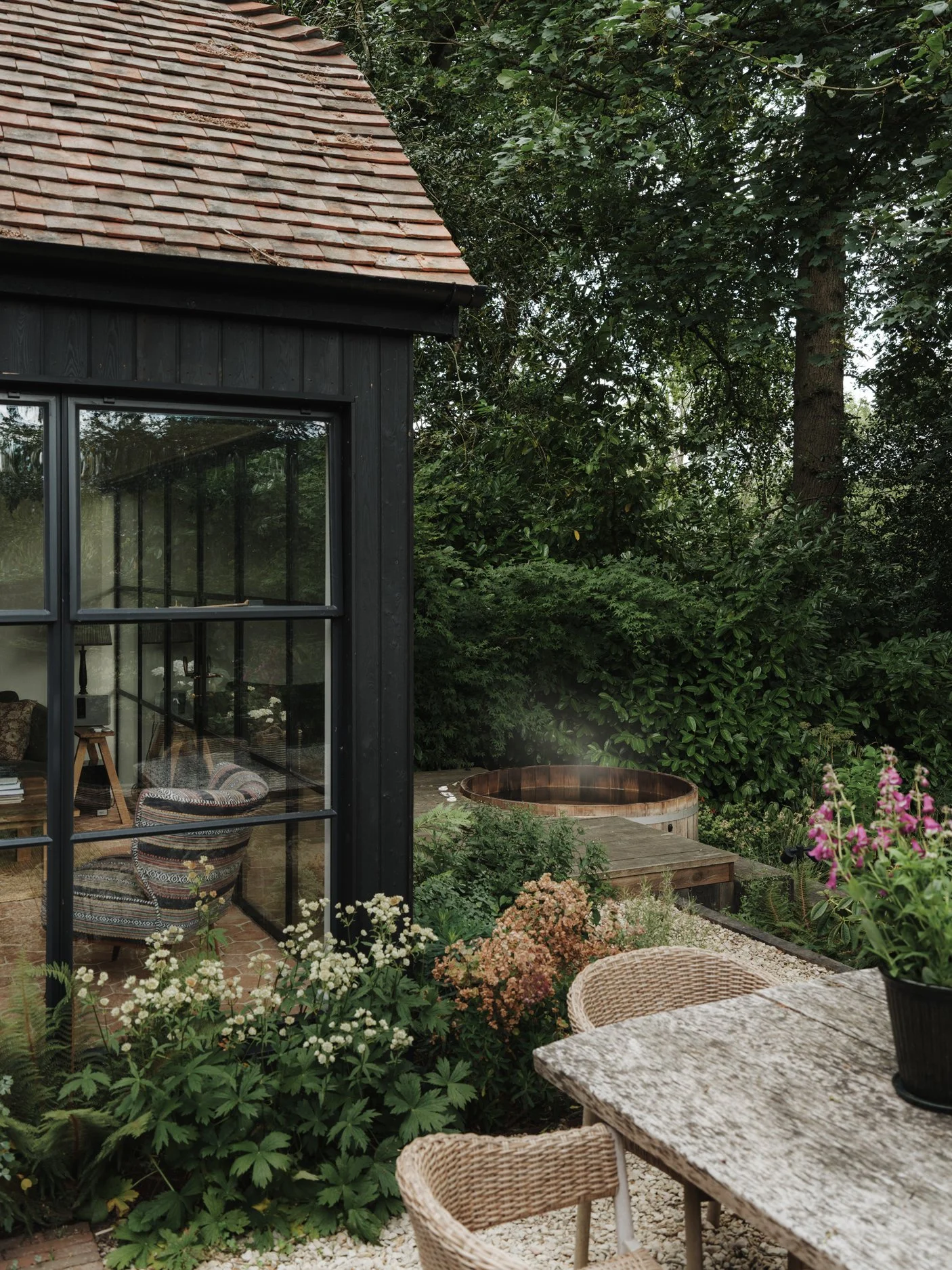 An outdoor garden area with a black wooden house that has a large window. Inside the house, a cozy seating area with patterned cushions is visible. Outside, there are various plants and shrubs, a weathered stone table with wicker chairs, and a large potted plant on the table. In the background, there is a wooden hot tub and tall green trees surrounding the area.