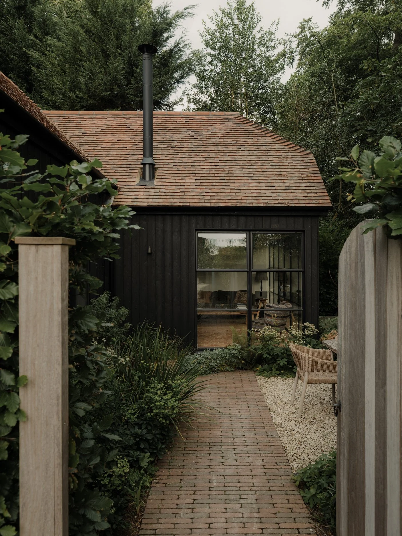 A small, black wooden house with a sloped roof and a large window looks into the living room. There is a brick pathway leading to the entrance, surrounded by greenery and garden plants. Two chairs and a small table are outside the house.
