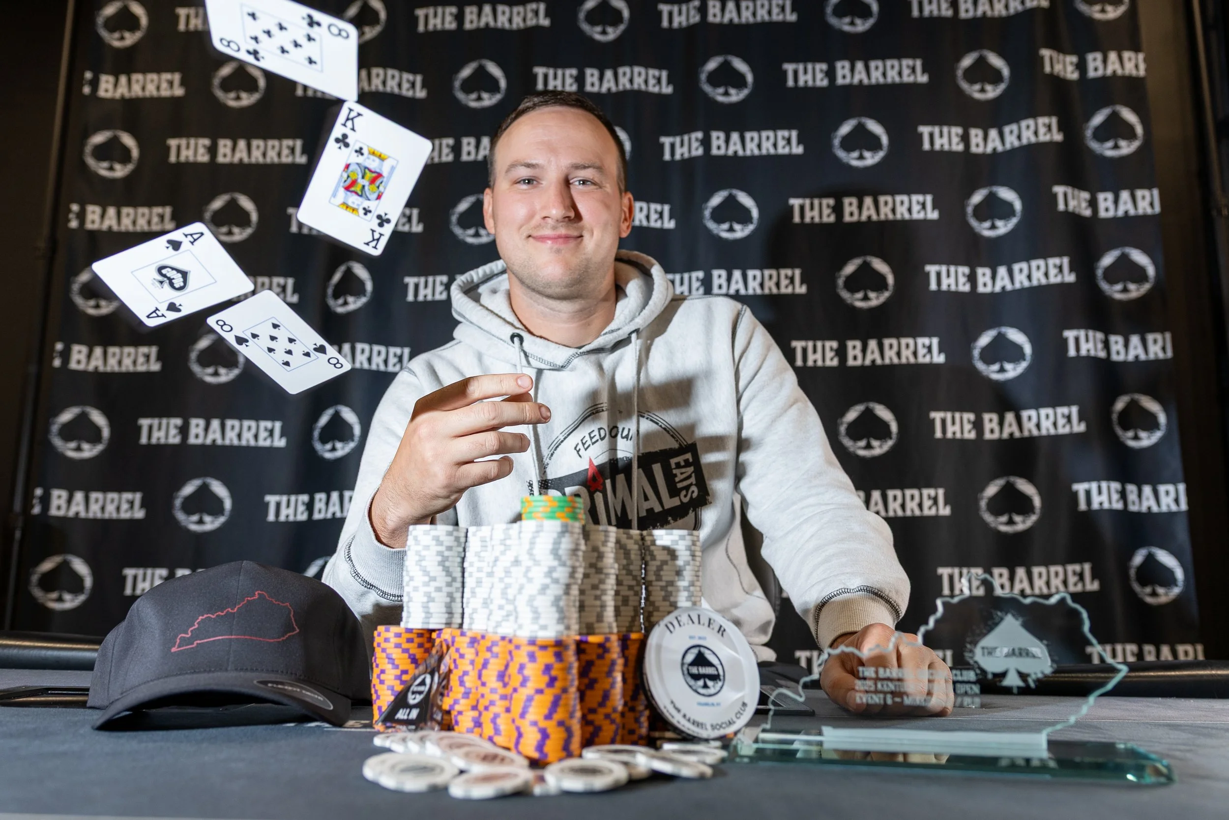 A man smiling at a poker table with poker chips, a hat, and playing cards, with a backdrop reading 'The Barrel'.