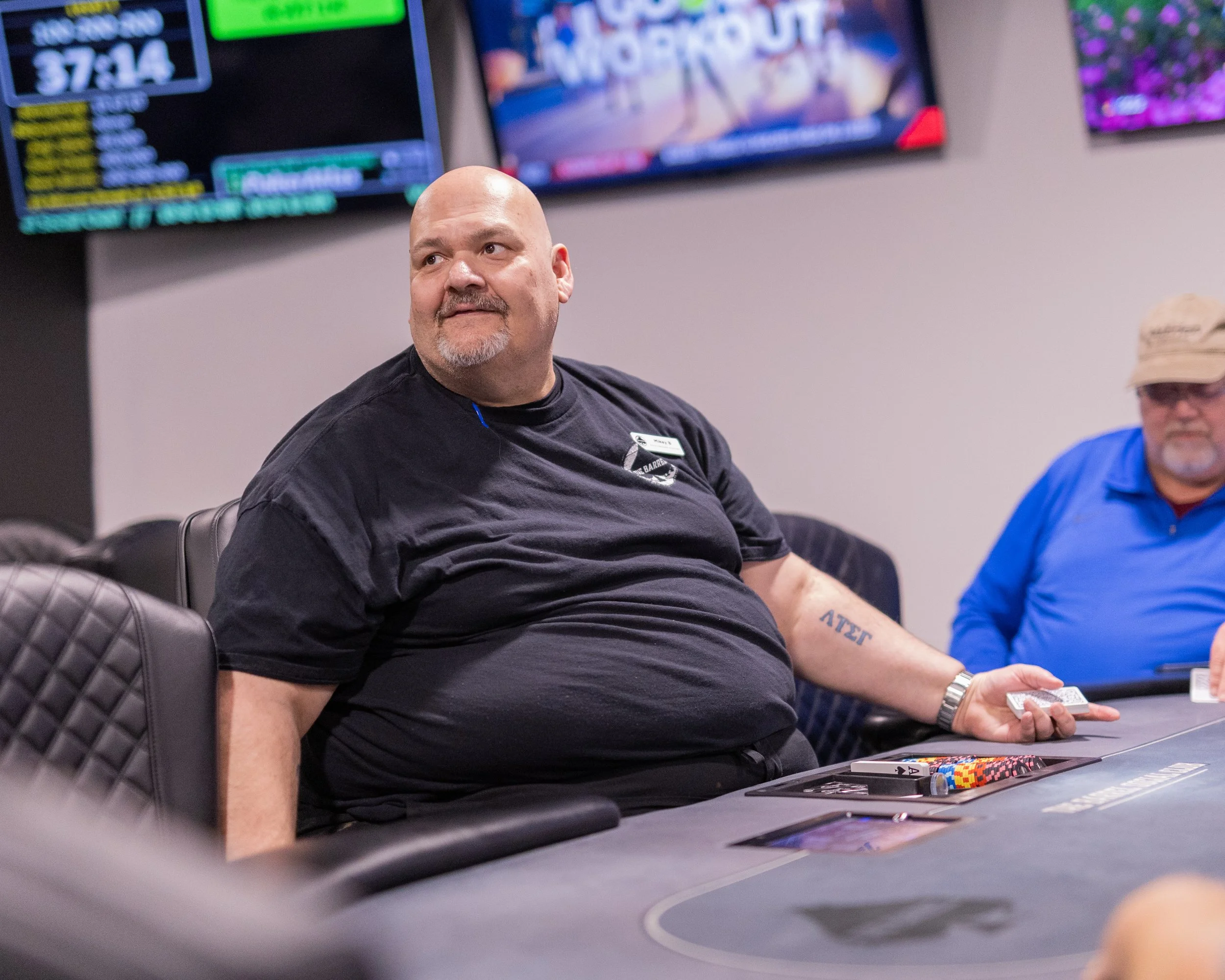 A man with a shaved head and beard, wearing a black shirt, sits at a poker table with poker chips, holding cards, and watching the game.