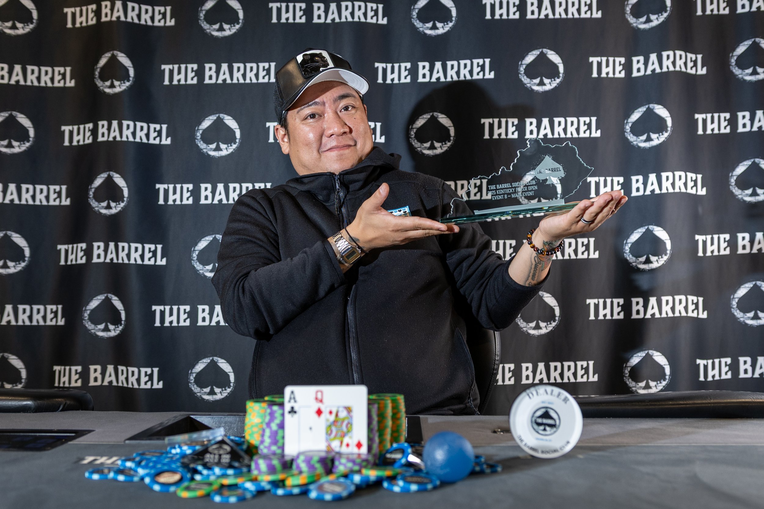 A man holding a playing card and an award plaque at a poker tournament, with poker chips and playing cards on the table, and a backdrop with 'The Barrel' logo.