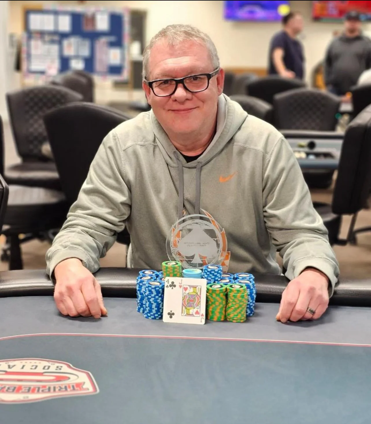 A man with glasses and a gray Nike hoodie sitting at a poker table, smiling, with a pile of poker chips and two playing cards in front of him. The two cards are a 4 of clubs and a Jack of hearts. The background shows other players and a bulletin board.