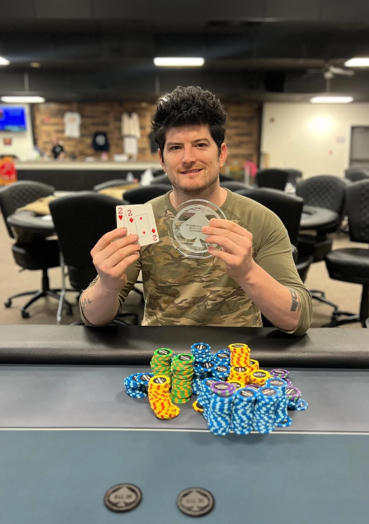 A man with dark hair and a camouflage shirt is sitting at a poker table, holding two two of diamonds playing cards in his left hand and a glass award in his right hand. There are stacks of colorful poker chips in front of him.