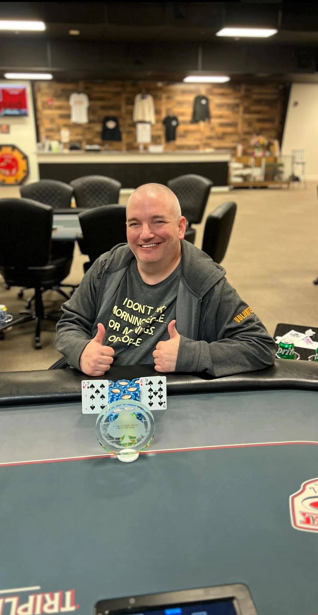 A man smiling and giving two thumbs up while seated at a poker table with playing cards and poker chips in front of him.