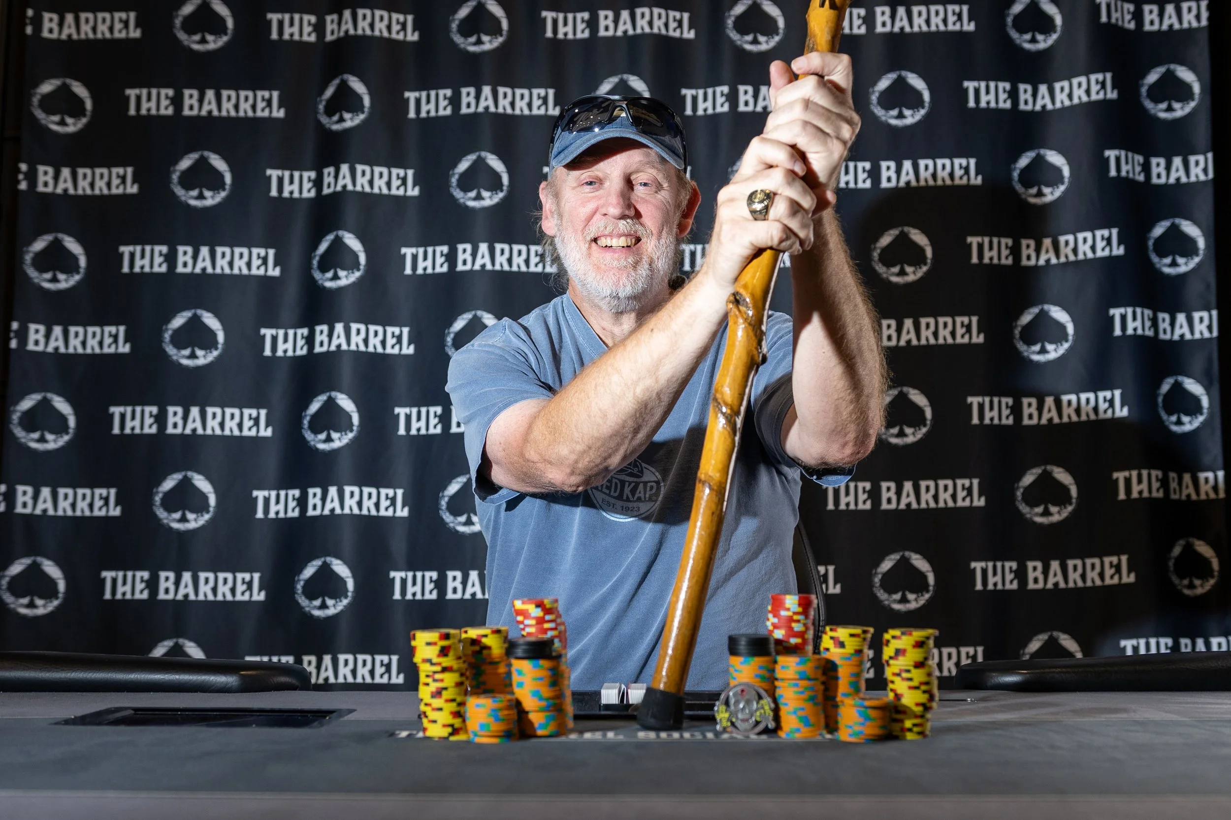 A smiling man with a beard and cap playing poker at a casino, with stacks of poker chips in front of him, and a backdrop with the text 'The Barrel'.