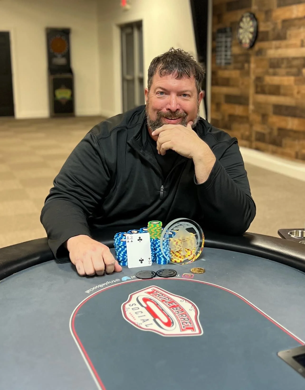 Man smiling at a poker table with chips, playing cards, coins, and a poker chip display in front of him.