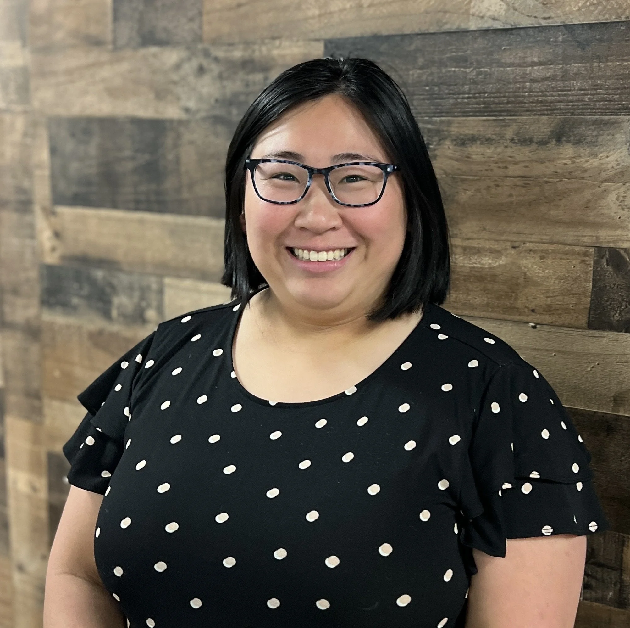 A woman with black hair, glasses, and a black polka-dot shirt smiling in front of a wooden wall.