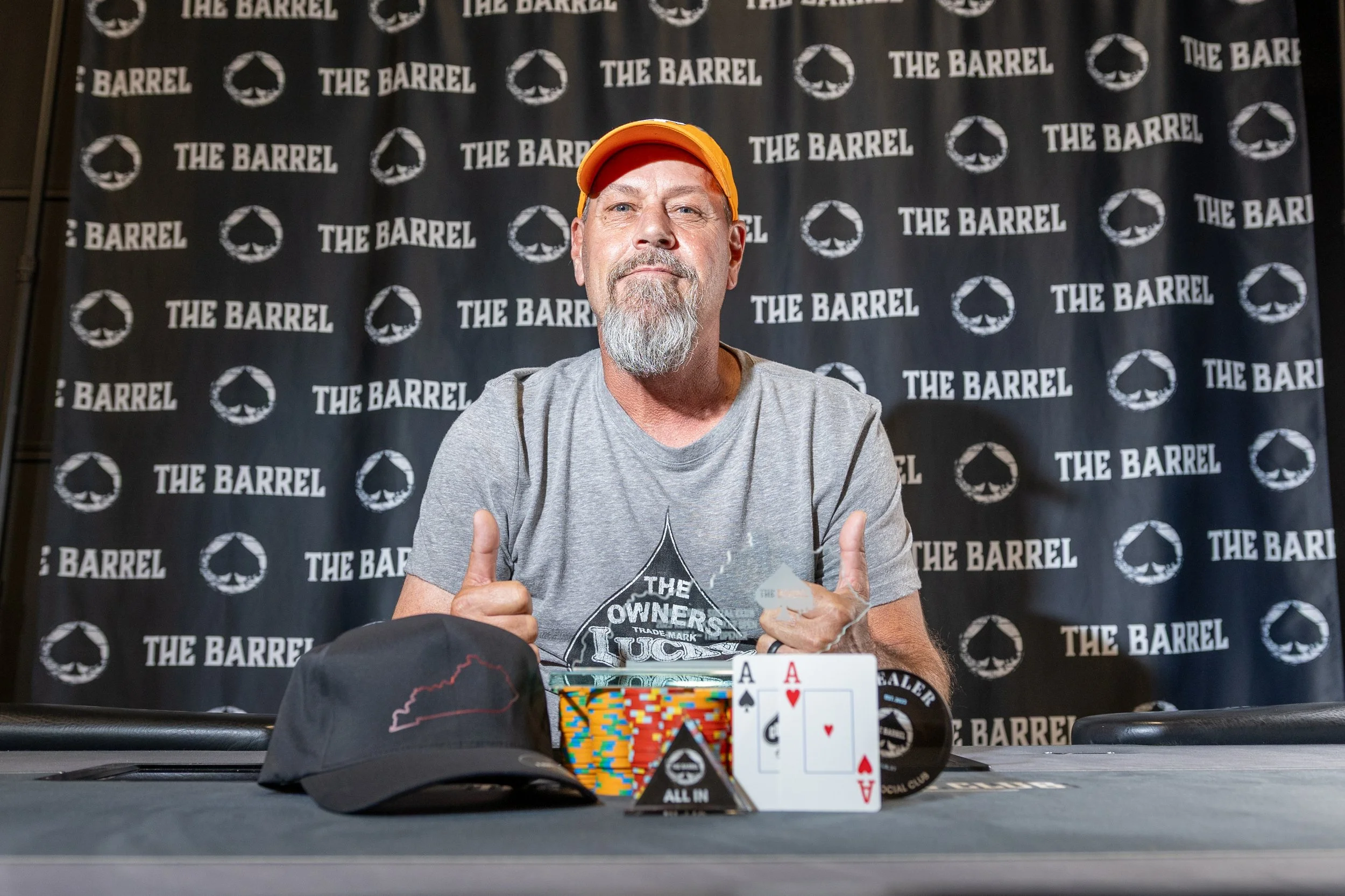 A man with a beard and mustache wearing an orange cap and gray t-shirt, giving two thumbs up, sitting in front of a backdrop with the text 'The Barrel'. In front of him, there are two playing cards, an ace of spades and an ace of hearts, along with poker chips and a black cap with a red outline of a state.