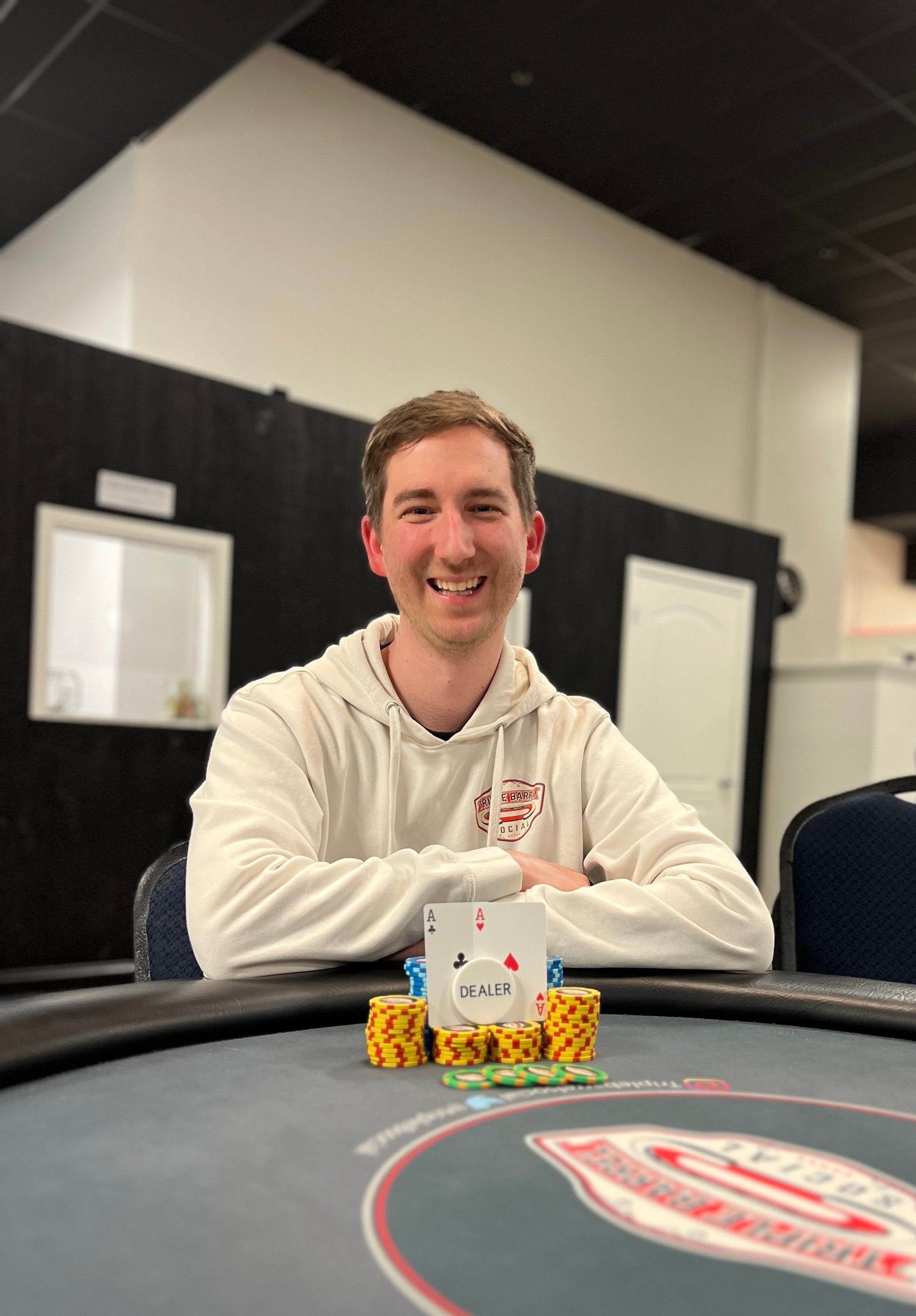 A young man with short brown hair, smiling, sitting at a poker table with stacks of chips and playing cards showing two aces, one of hearts and one of clubs, with a dealer button in front of him. He is wearing a white hoodie with a logo on it, in a room with black walls and white doors.