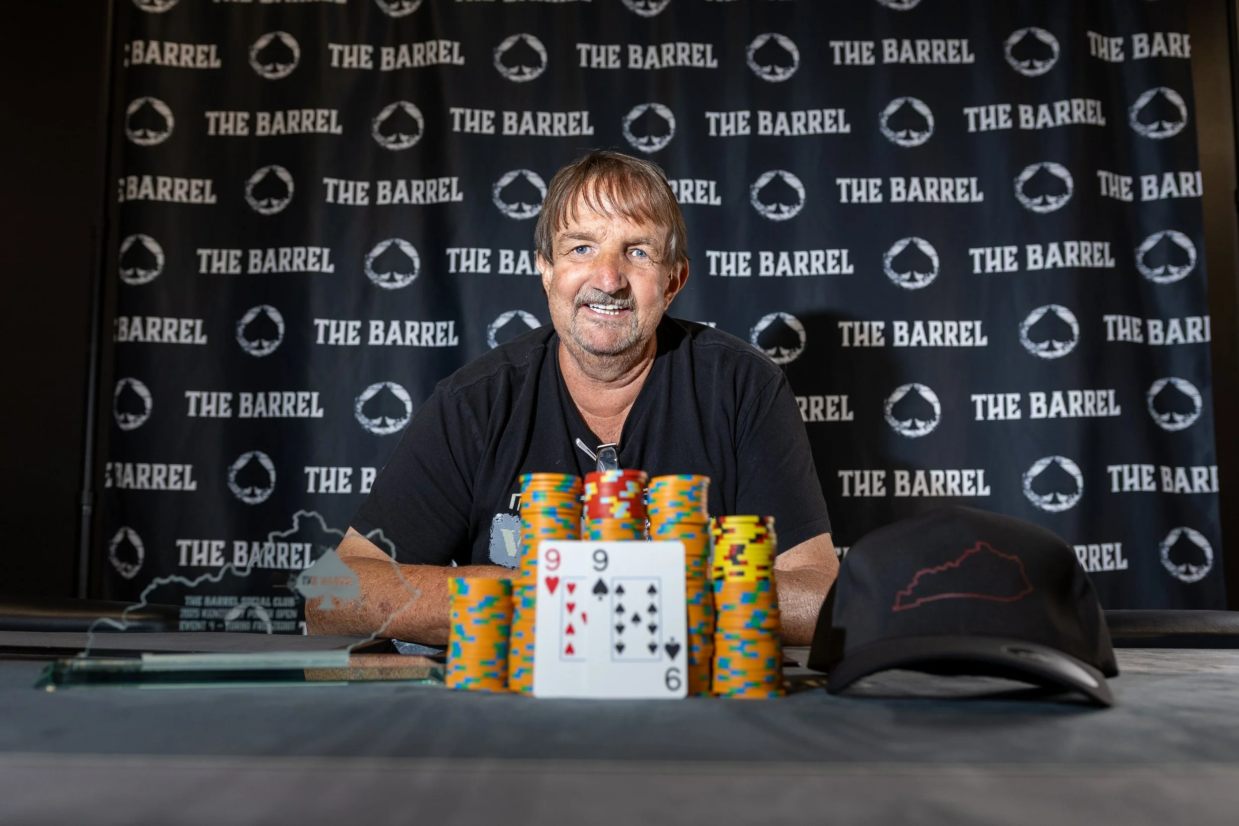A man with light brown hair, blue eyes, and a goatee, smiling at a poker tournament. He is seated at a table with poker chips and cards, including the 9 of hearts, 9 of spades, 6 of diamonds, and 10 of clubs. The background features a black banner with the logos and name of the poker venue, 'The Barrel'.