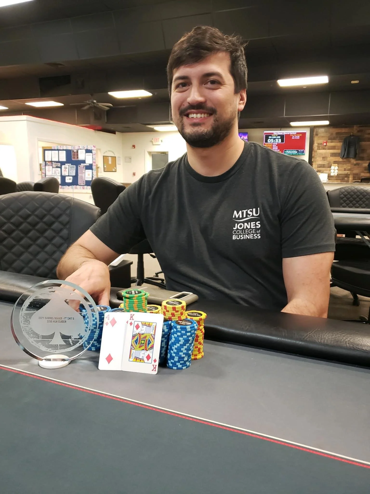 A man smiling at a poker table with poker chips, playing cards, and a trophy in front of him in a poker room. The man is wearing a black T-shirt with the words 'MTSU Jones College of Business' on it.