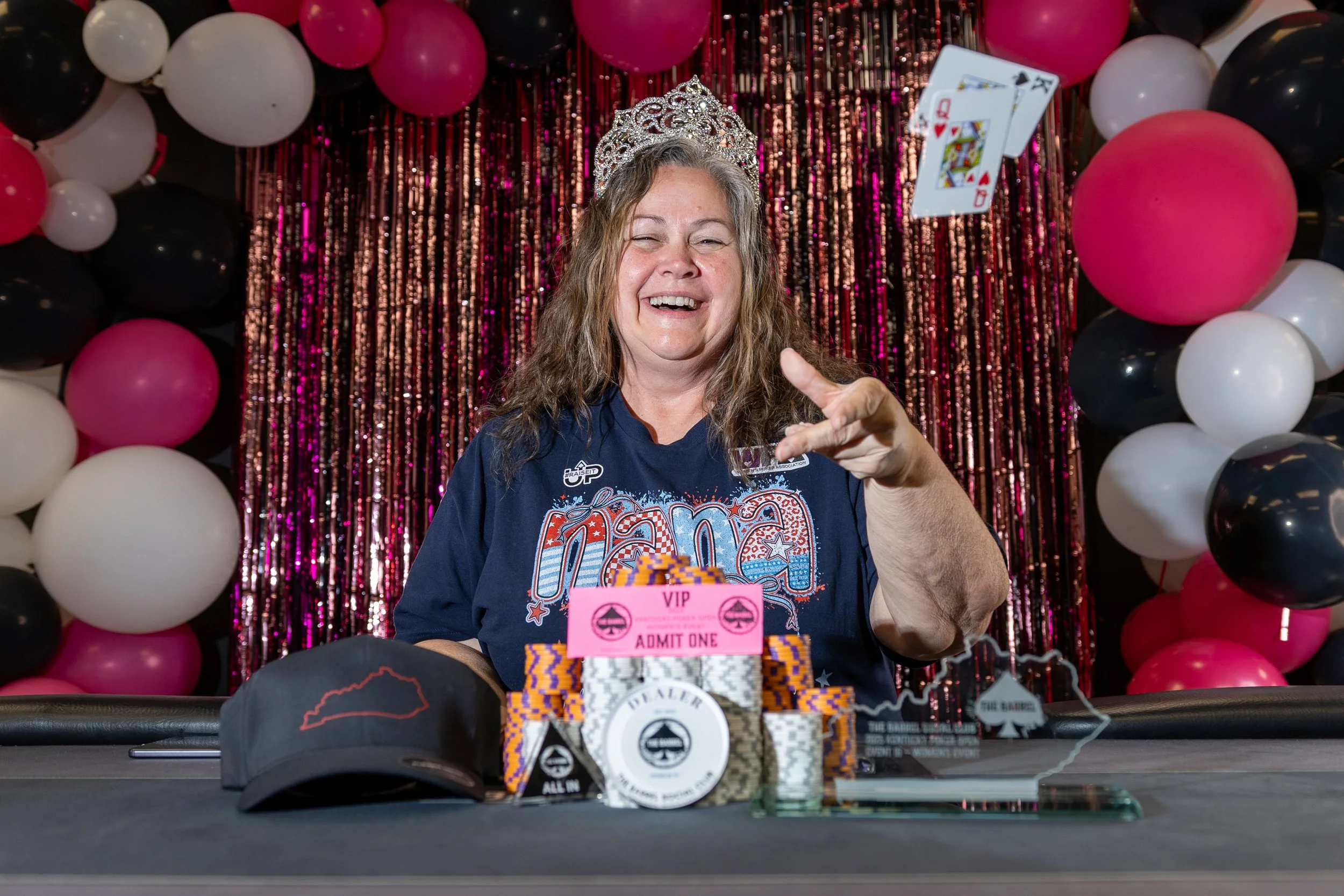 Woman wearing a tiara, smiling, and pointing at the camera behind a table with poker chips, hats, and a pink VIP ticket, with a backdrop of pink, white, and black balloons and metallic streamers.