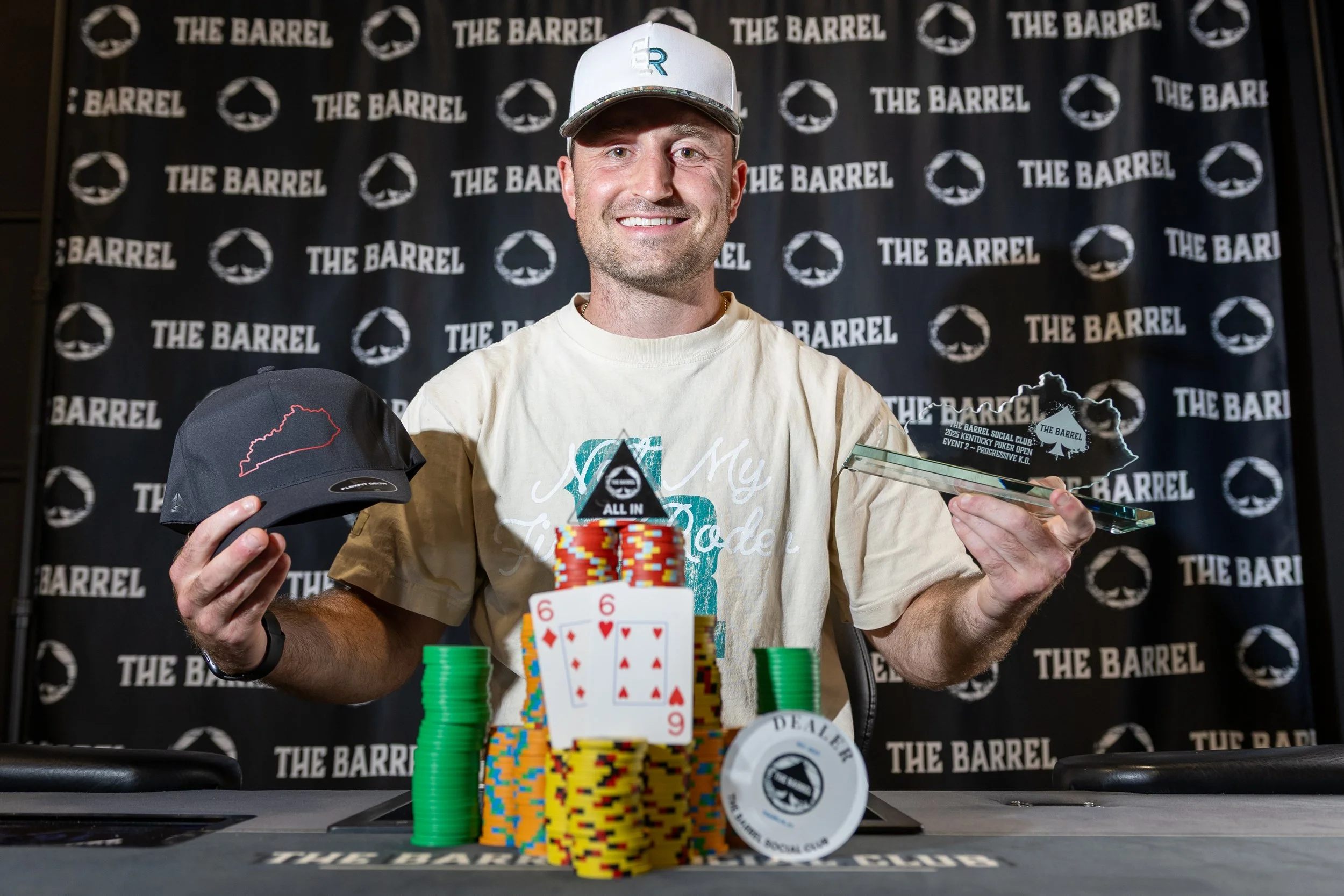 A man smiling at a poker tournament, holding a poker hat and a trophy, with poker chips and playing cards on the table, and a backdrop reading 'The Barrel'.