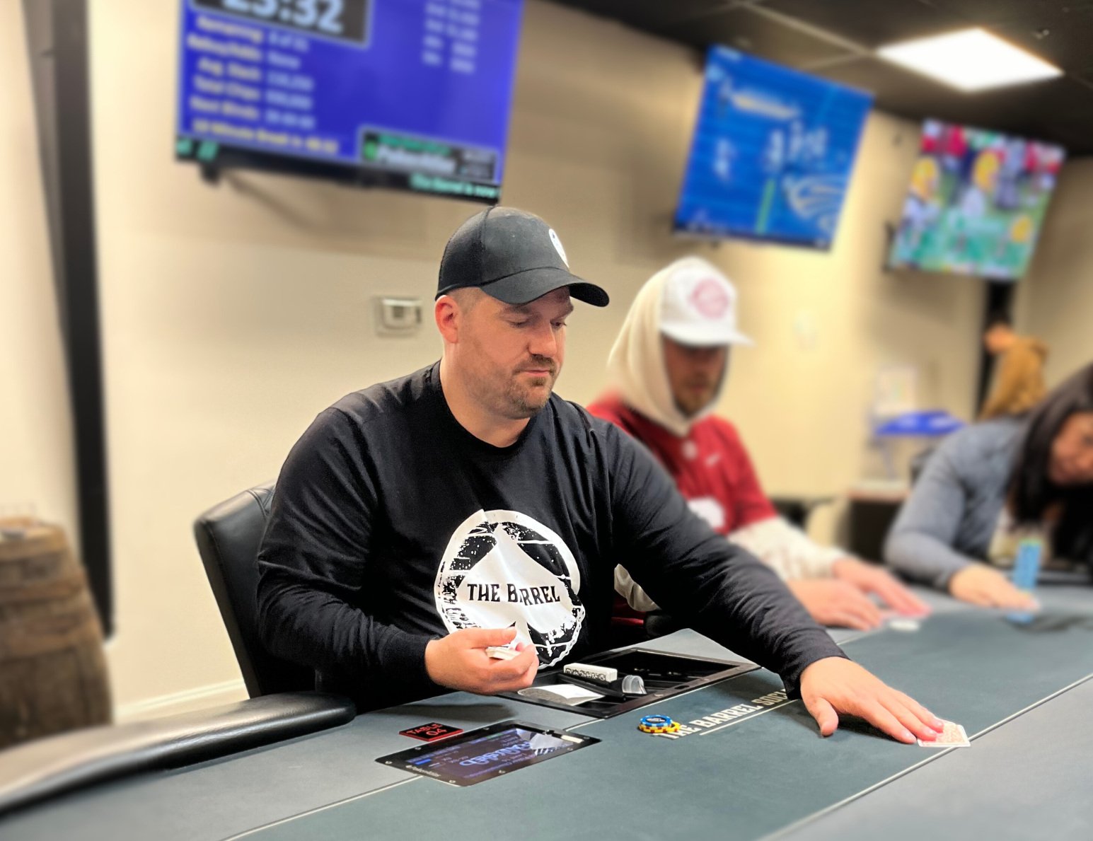 A man with a black cap and black long-sleeve shirt sitting at a poker table, holding playing cards, with poker chips in front of him. Other people are seated nearby, and multiple screens are hanging on the wall behind.