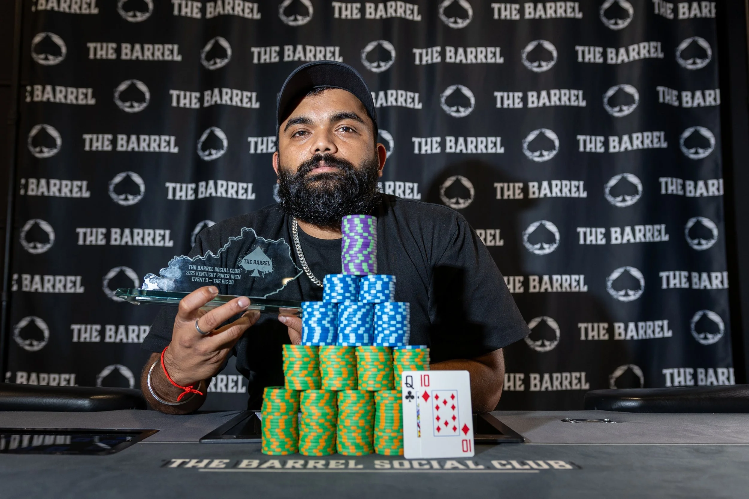 A man with a beard and wearing a cap sits behind a poker table, holding a wooden plaque, with poker chips and two playing cards (Queen of Clubs and Ten of Diamonds) in front of him, against a backdrop with the words 'The Barrel' and a spade symbol.