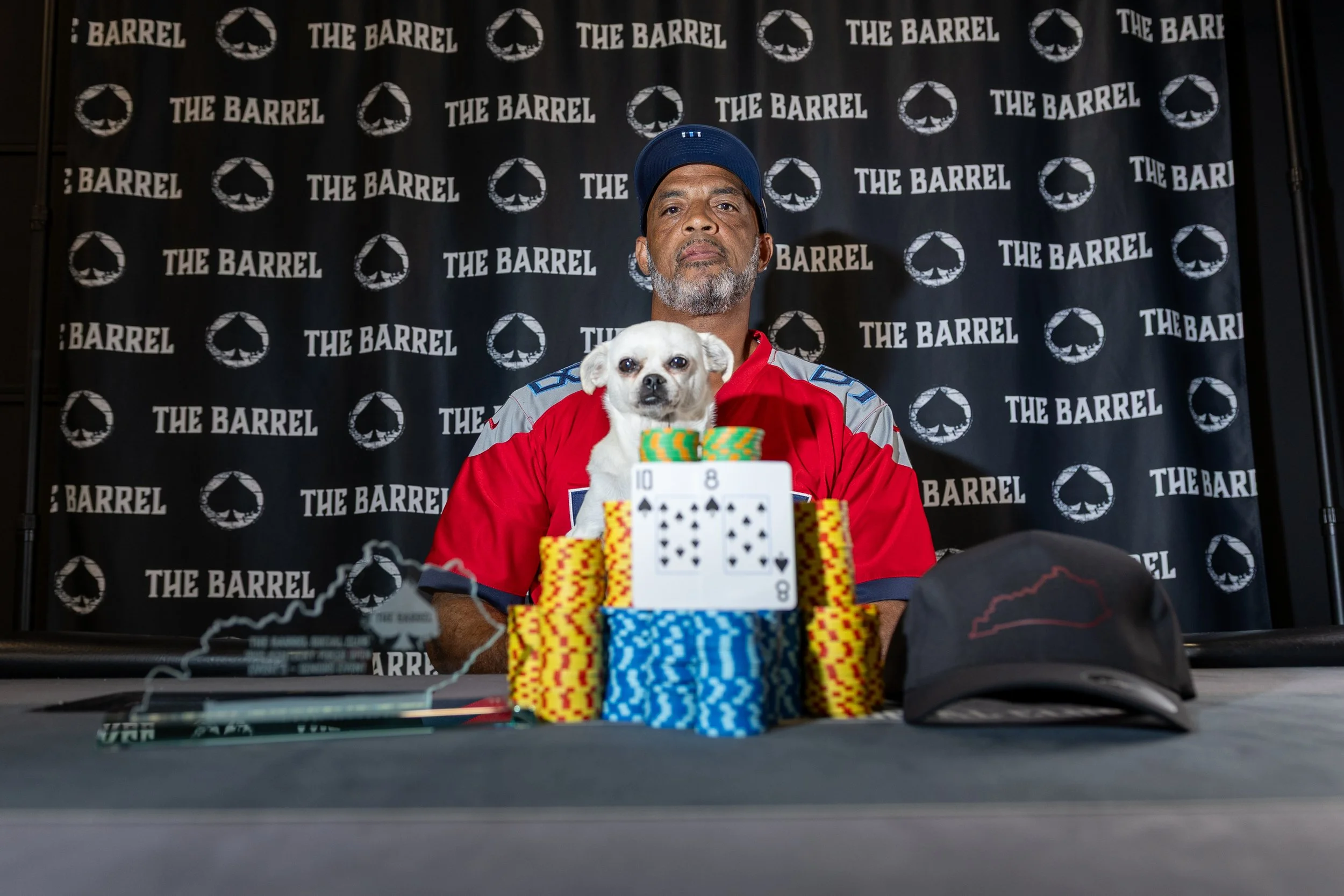 A man with a gray beard and a blue cap, sitting at a poker table with a small dog sitting on his lap. The background features a black backdrop with the logo and name "The Barrel" repeated across it.