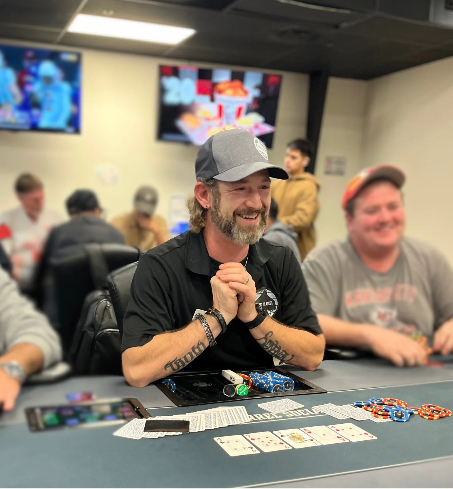 A man with a beard, wearing a grey and black cap, smiling with his hands clasped in front of him, sitting at a poker table with playing cards, poker chips, and a smartphone in front of him. There are other people in the background at the same table.