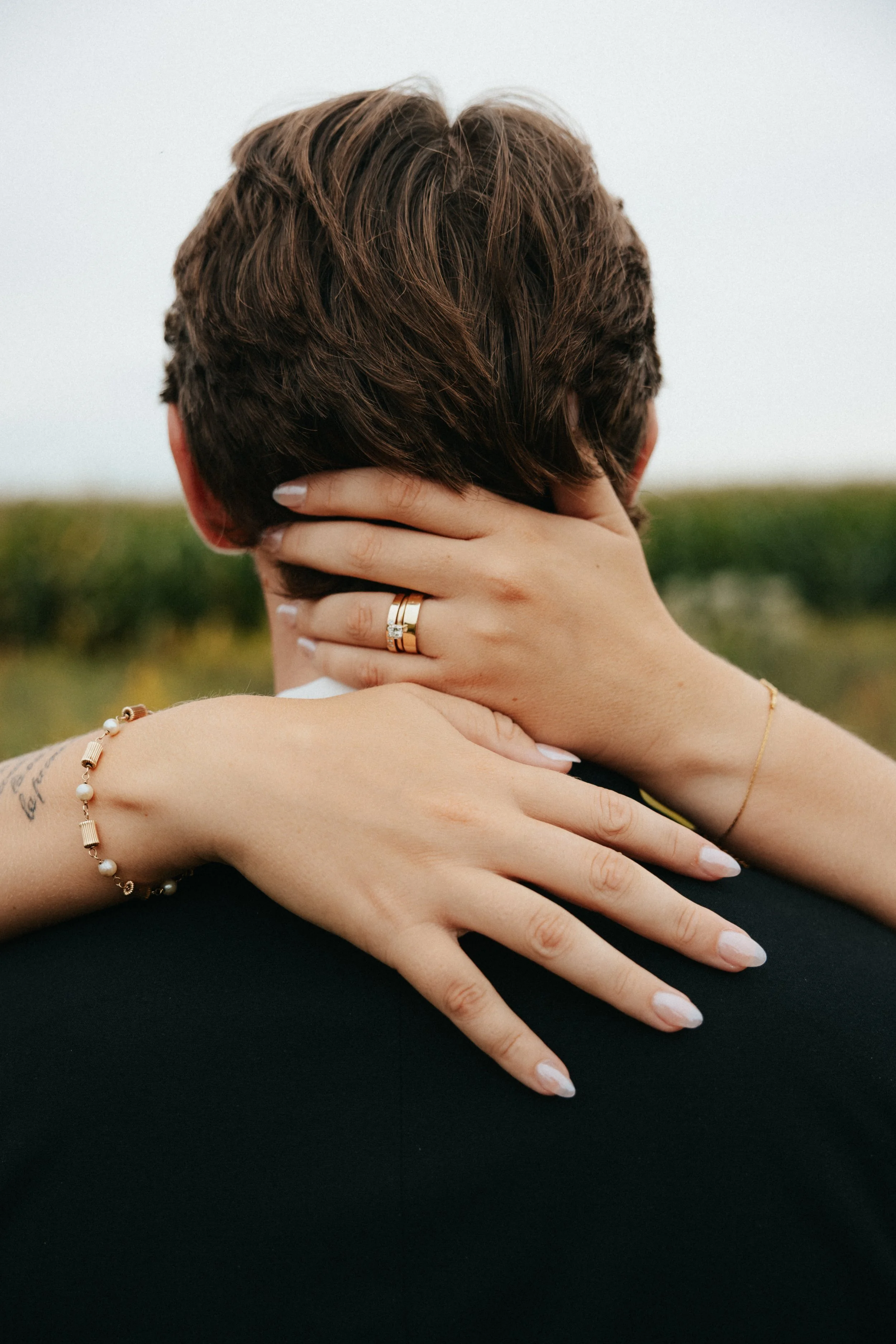 Person with short brown hair and light skin holding their neck with hands, wearing a pearl bracelet, a ring, and a thin bracelet, with a blurred outdoor background.