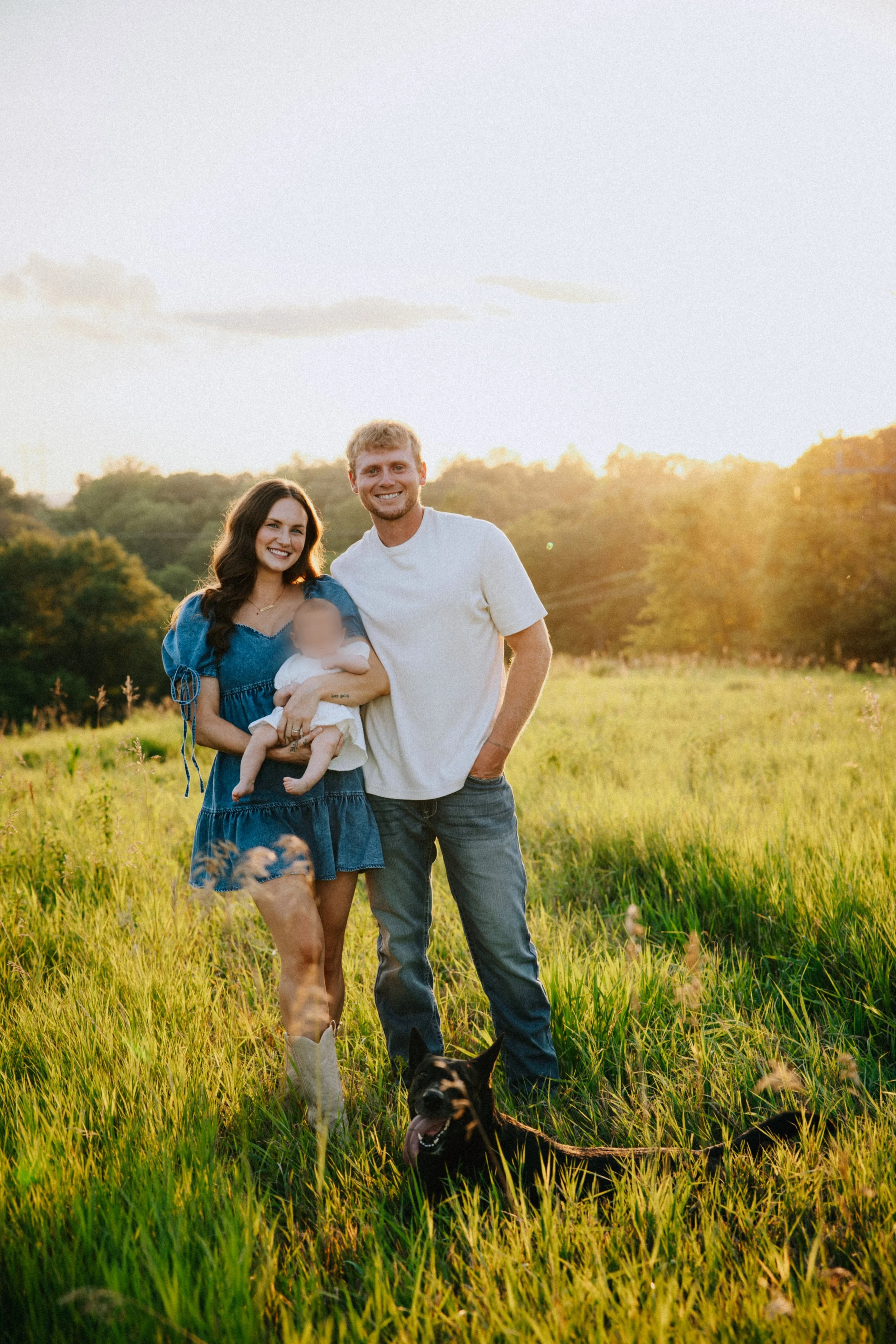 A happy couple and their dog standing in a grassy field during sunset. The woman is holding a baby, and the man is standing next to her. The dog, a black and tan breed, is sitting in front of them with its tongue out.