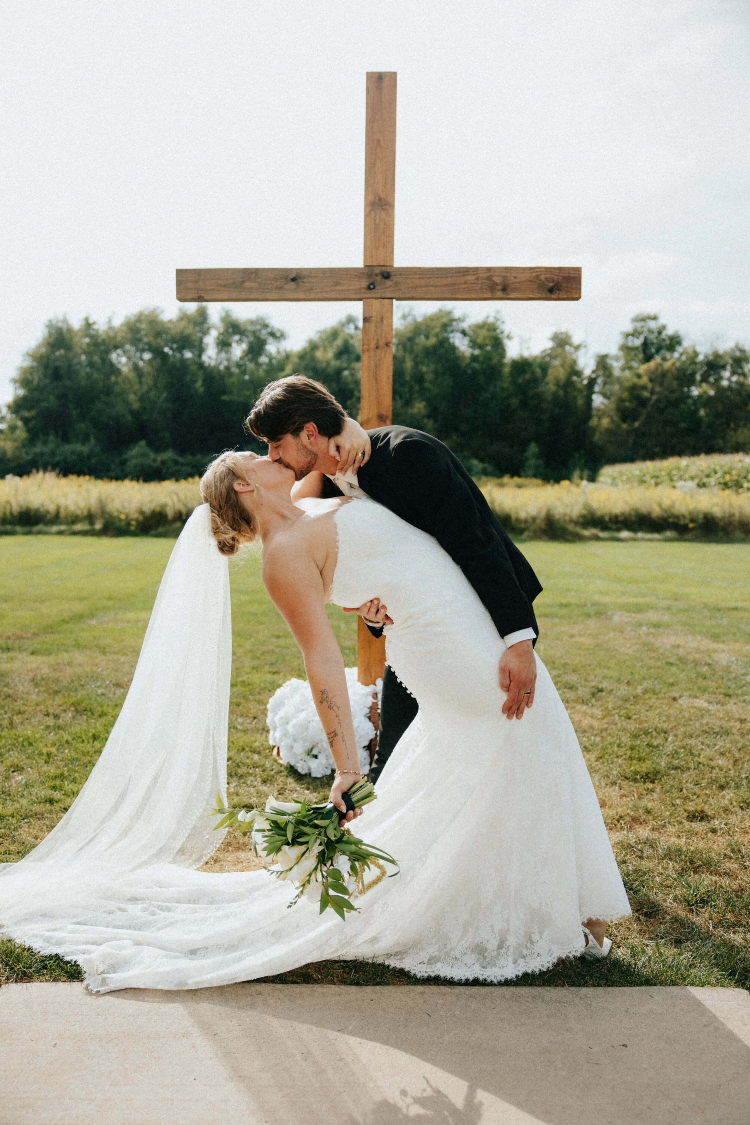 A bride and groom kiss outdoors during their wedding ceremony, with a large wooden cross in the background.