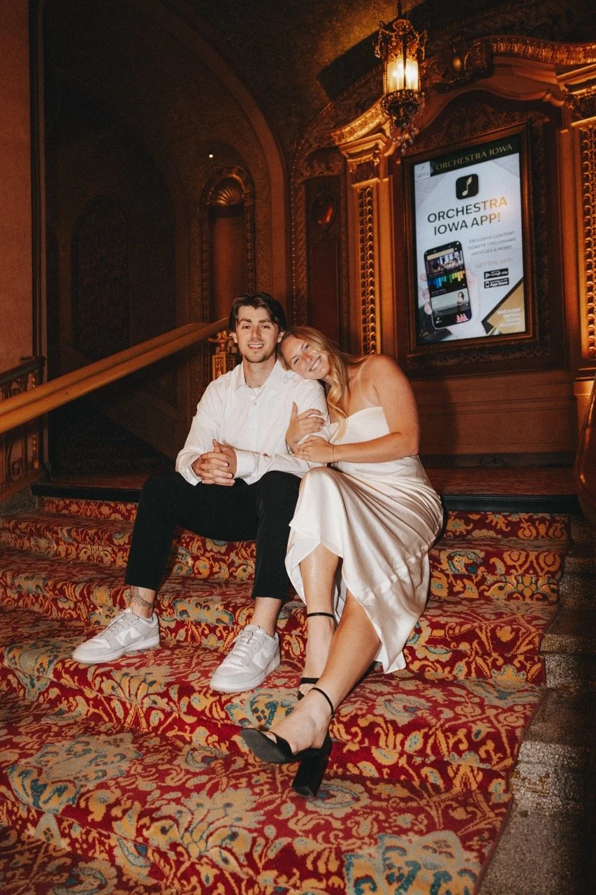 A man and woman sitting on ornate red carpeted stairs, likely in a theater or concert hall, smiling and posing for the camera. The woman is leaning her head on the man's shoulder, wearing a white dress and black heels. The man is dressed in a white s