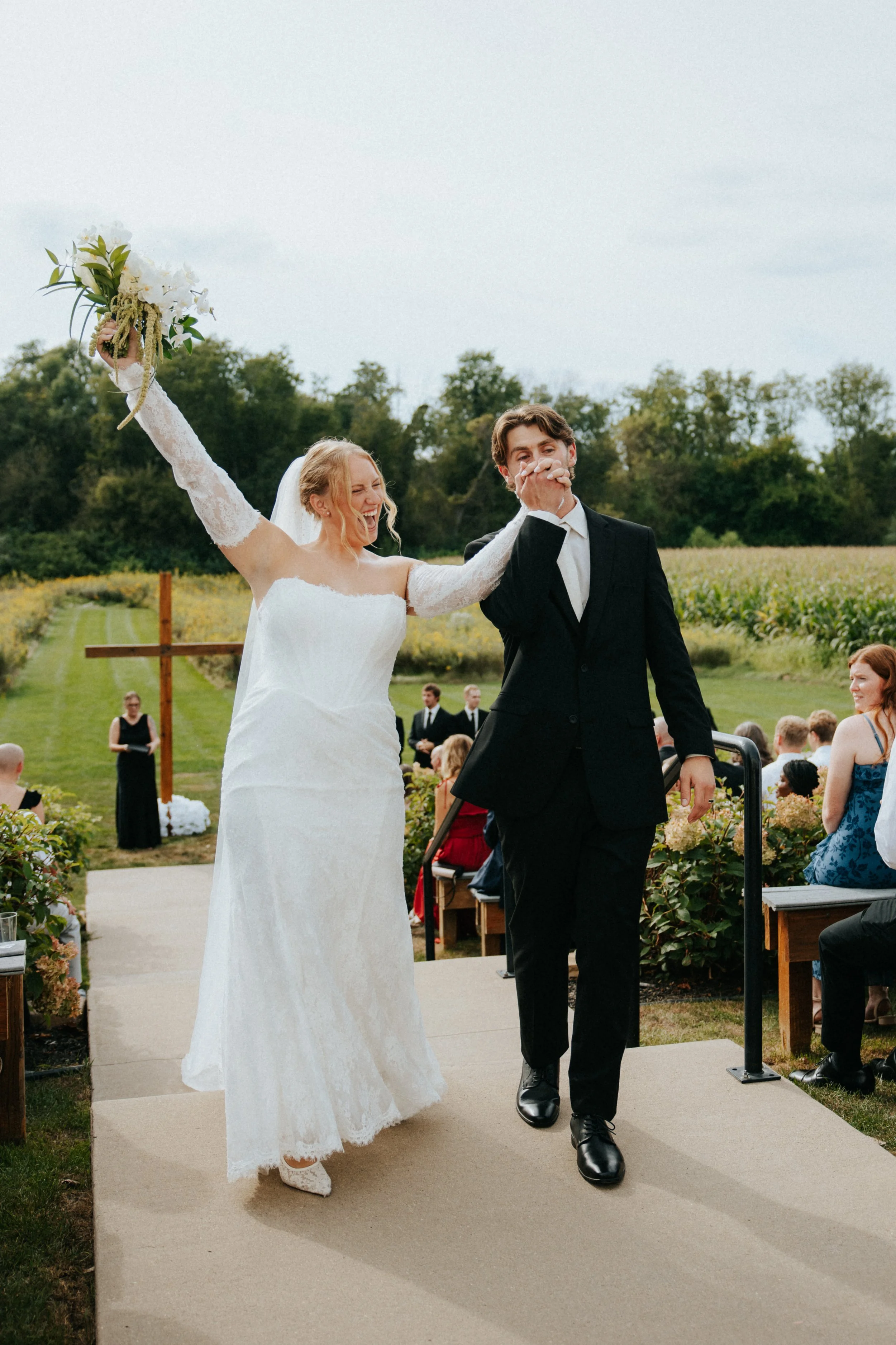A bride and groom celebrating outdoors, with the bride smiling and raising her bouquet, while the groom covers his mouth, at a wedding ceremony in a rural setting with guests seated nearby.