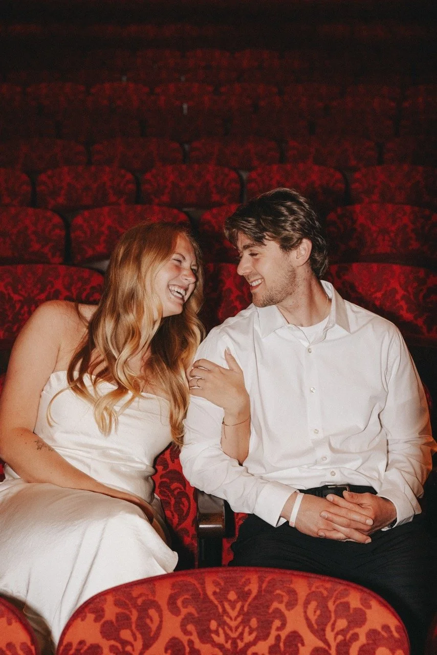 A smiling couple sitting close together in an empty theater with red velvet seats, sharing a joyful moment.