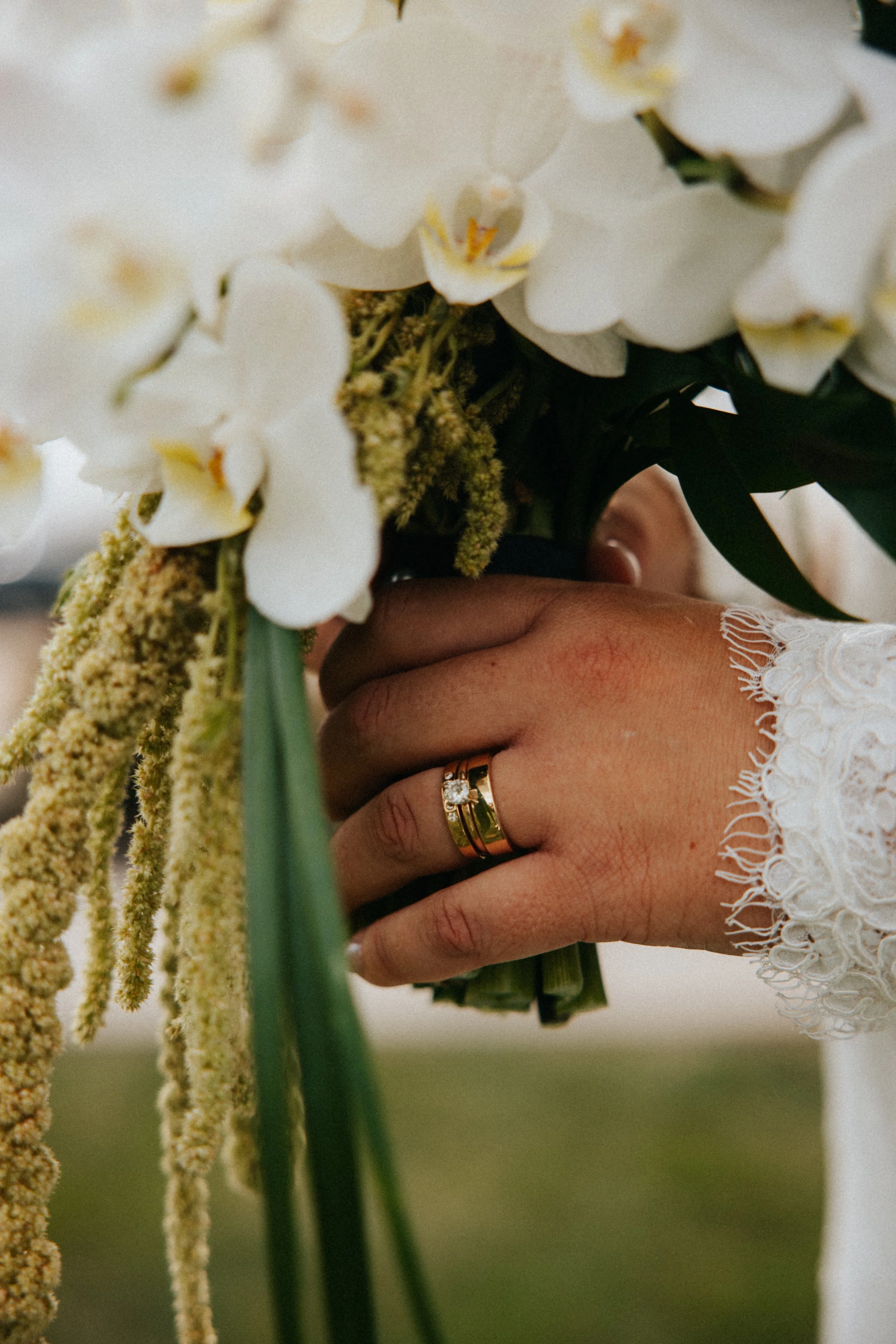 Close-up of a person's hand with wedding rings, holding a bouquet of white orchids and yellow flowers.