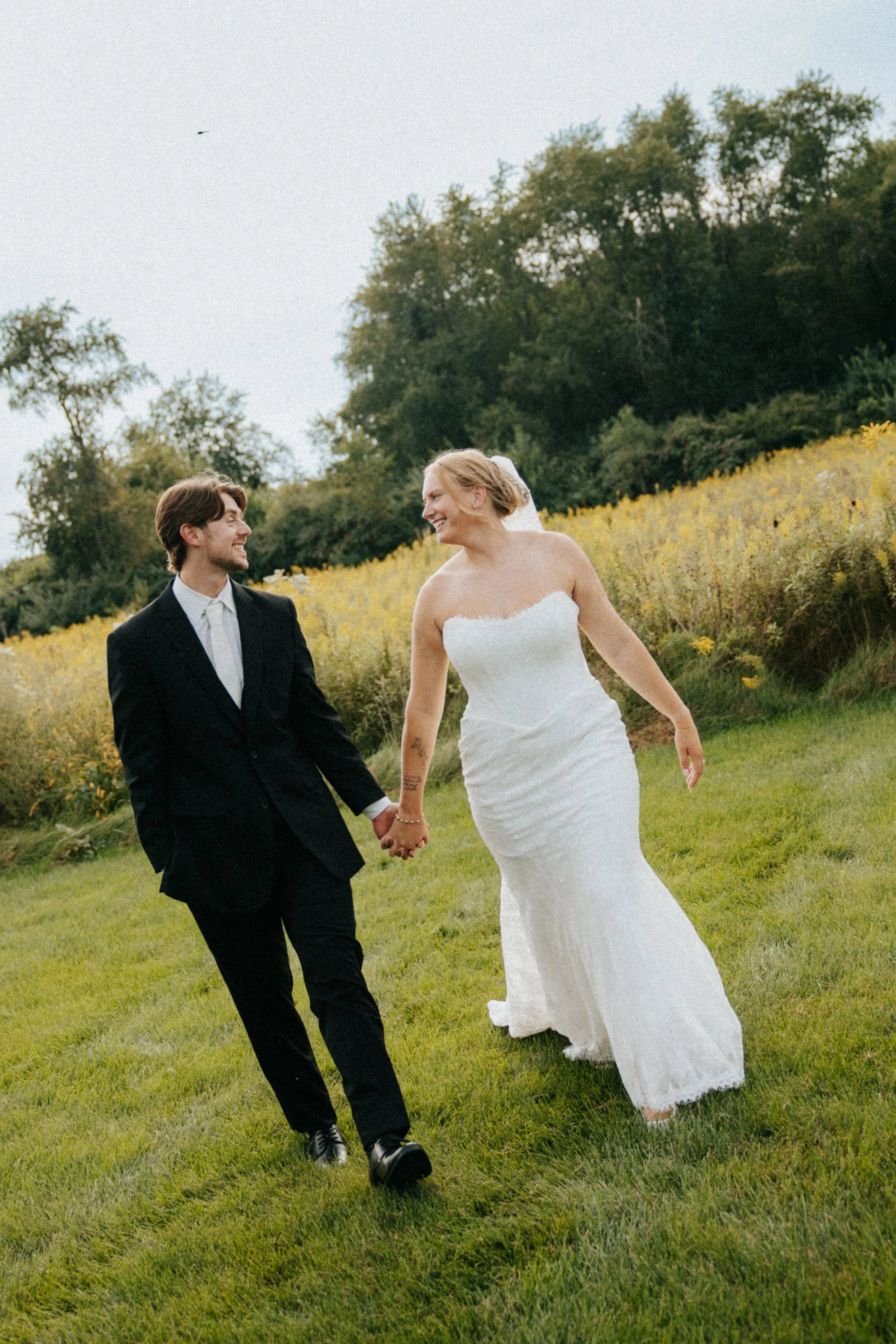 A couple in wedding attire walking hand in hand through a grassy field, with yellow wildflowers and trees in the background, smiling and looking at each other.