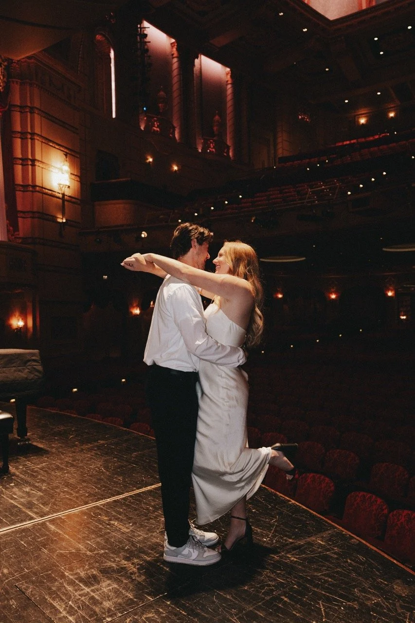 A couple dancing closely on a stage in an elegant theater with warm lighting and ornate architecture.