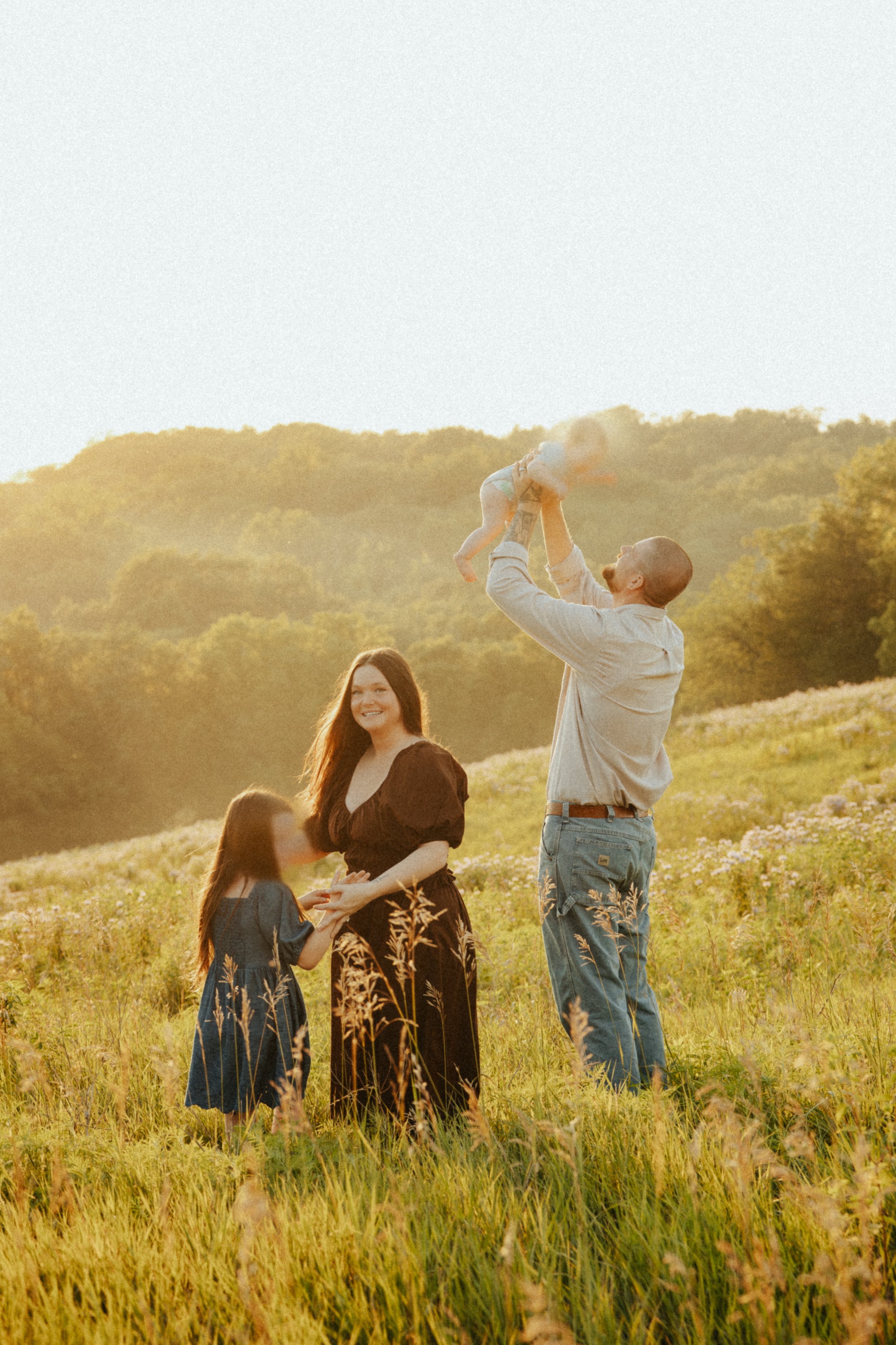 Family of five enjoying a sunny day in a field, with a man holding a baby up in the air, and smiling woman, girl, and boy standing nearby, with a green hill and trees in the background.
