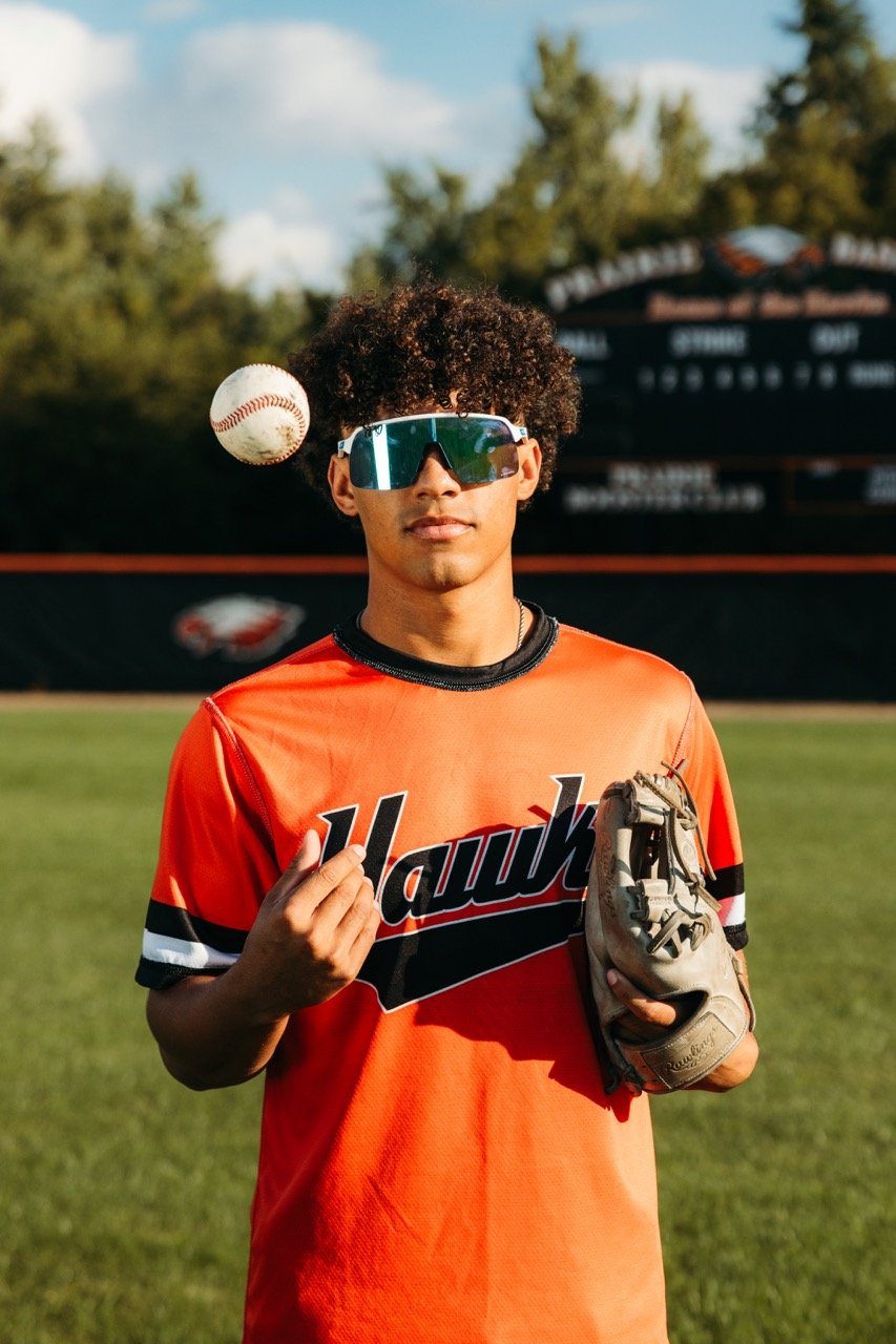A young male baseball player in an orange Hawkeyes jersey, wearing large sports sunglasses, holding a baseball glove in one hand, while making a hand gesture with the other, standing on a baseball field with a scoreboard and trees in the background.