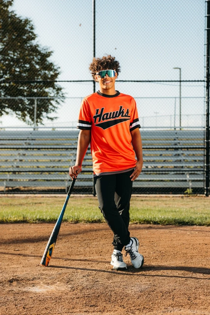 A young woman in sports gear standing on a baseball field, holding a baseball bat, wearing an orange jersey with 'Hawks' written on it, athletic pants, and sunglasses, smiling.