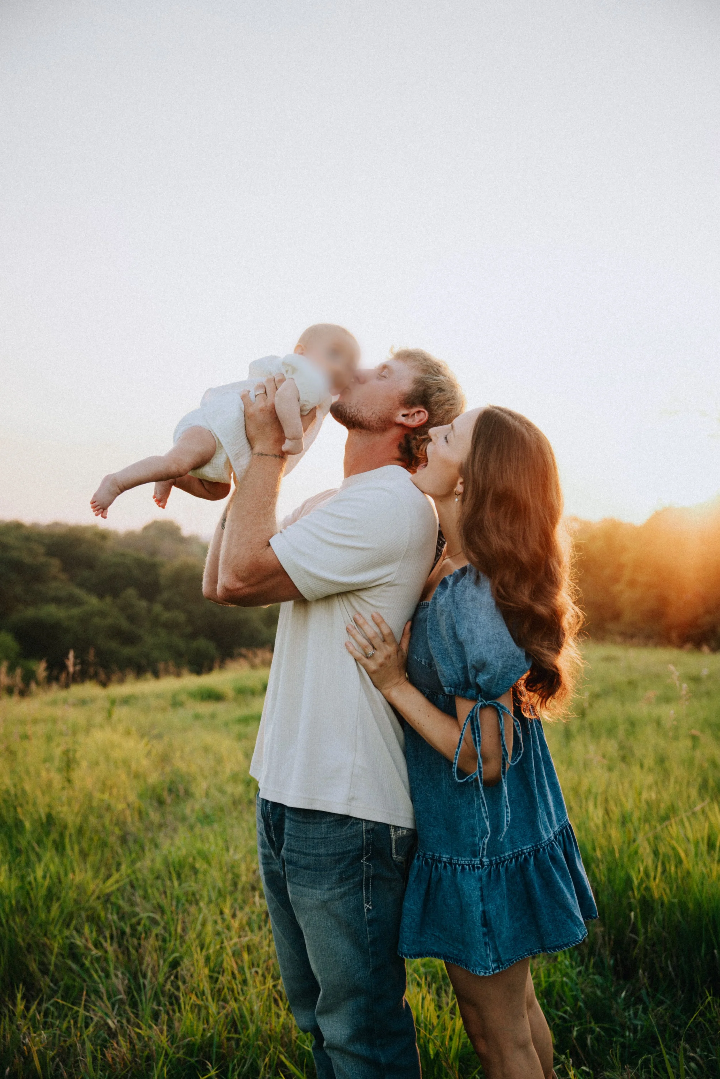A family of three in a grassy field during sunset. The father is holding and kissing a baby, while the mother embraces the father from behind. The sky is light with the setting sun, creating a warm, golden atmosphere.