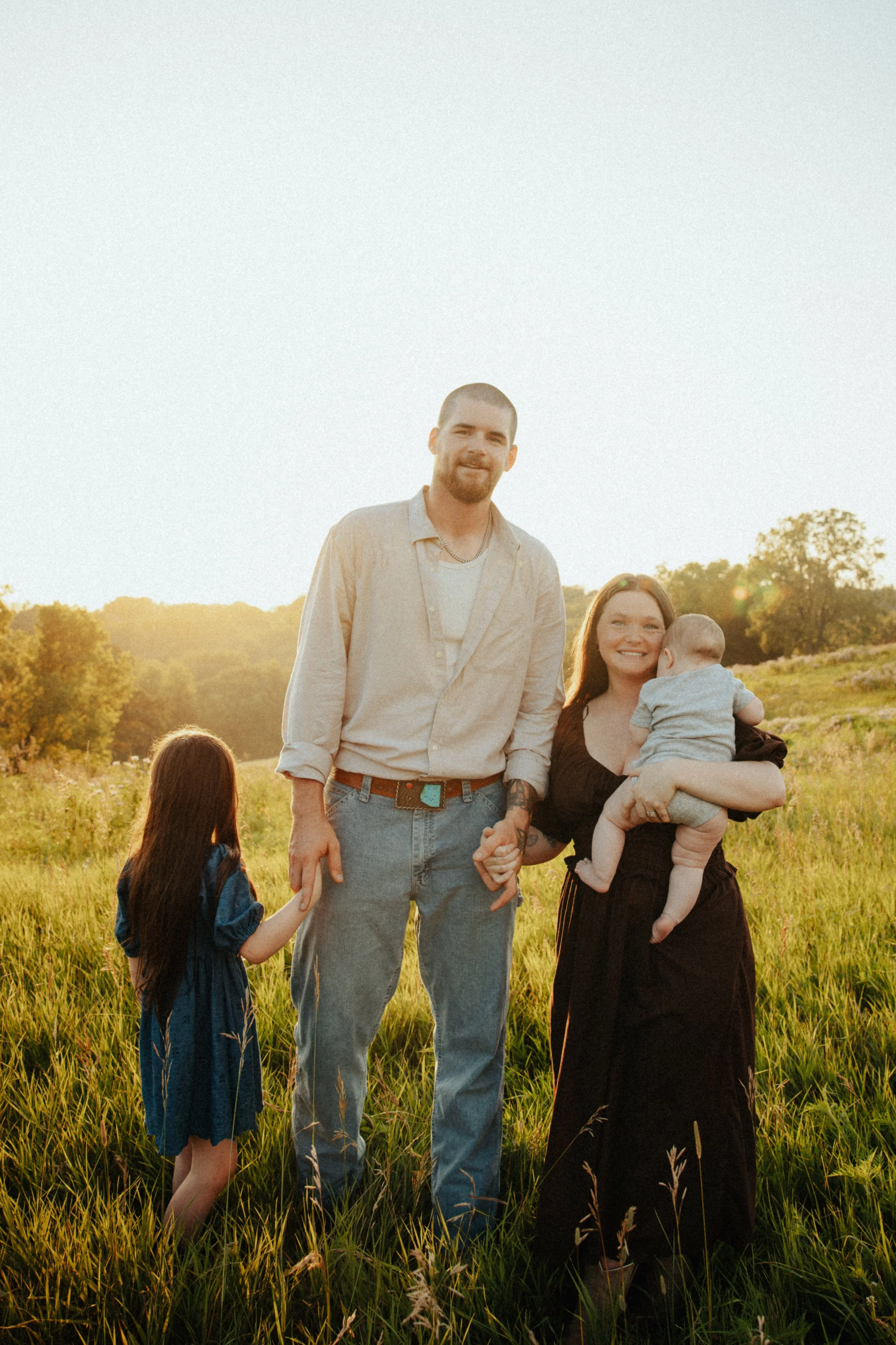 A family standing in a grassy field during sunset. A man with a beard in a beige shirt holds hands with a young girl. A woman is holding a baby, and another girl with long hair is facing away from the camera.