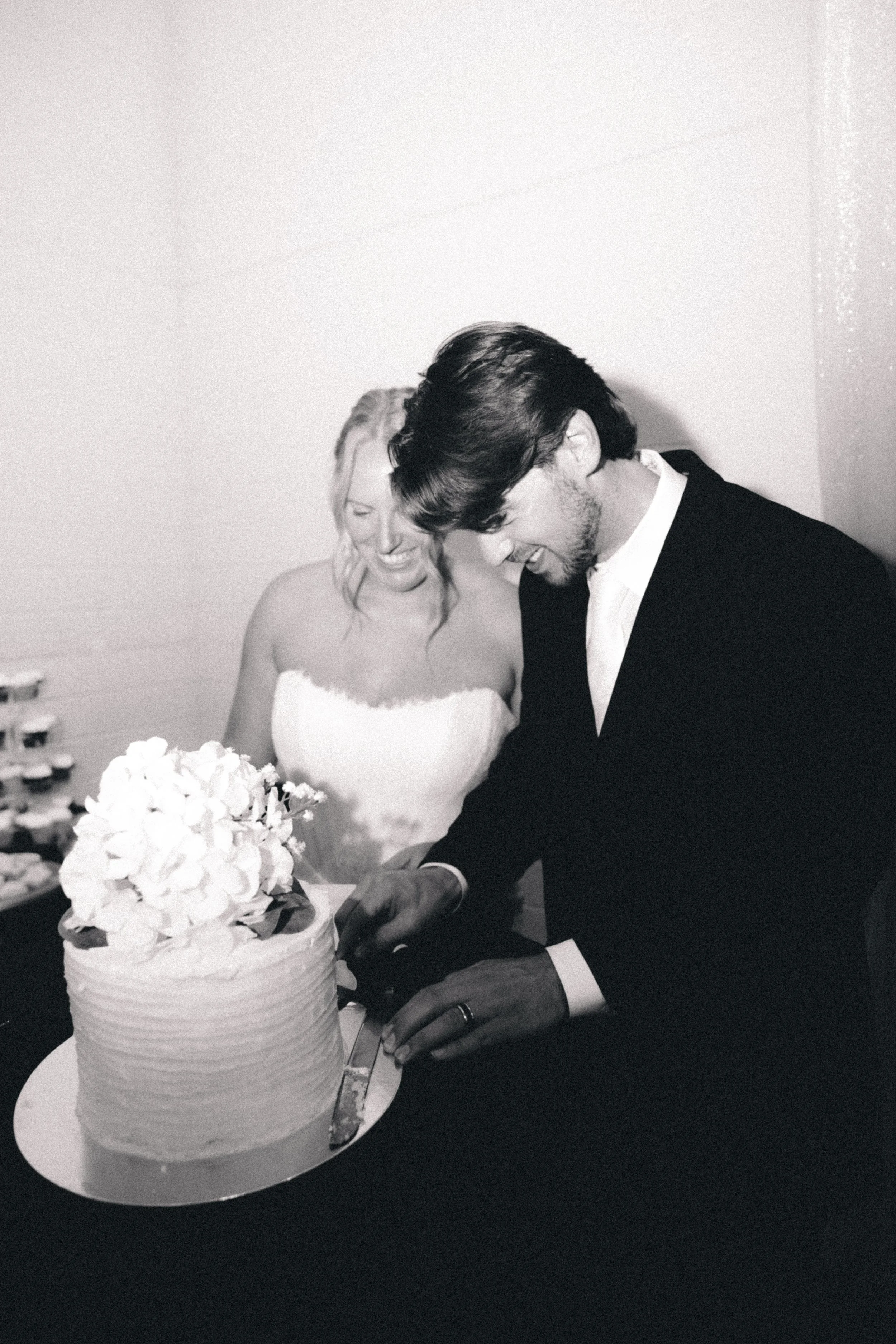 Black and white photo of a man and woman cutting wedding cake, woman in white dress, man in dark suit with tie, smiling.