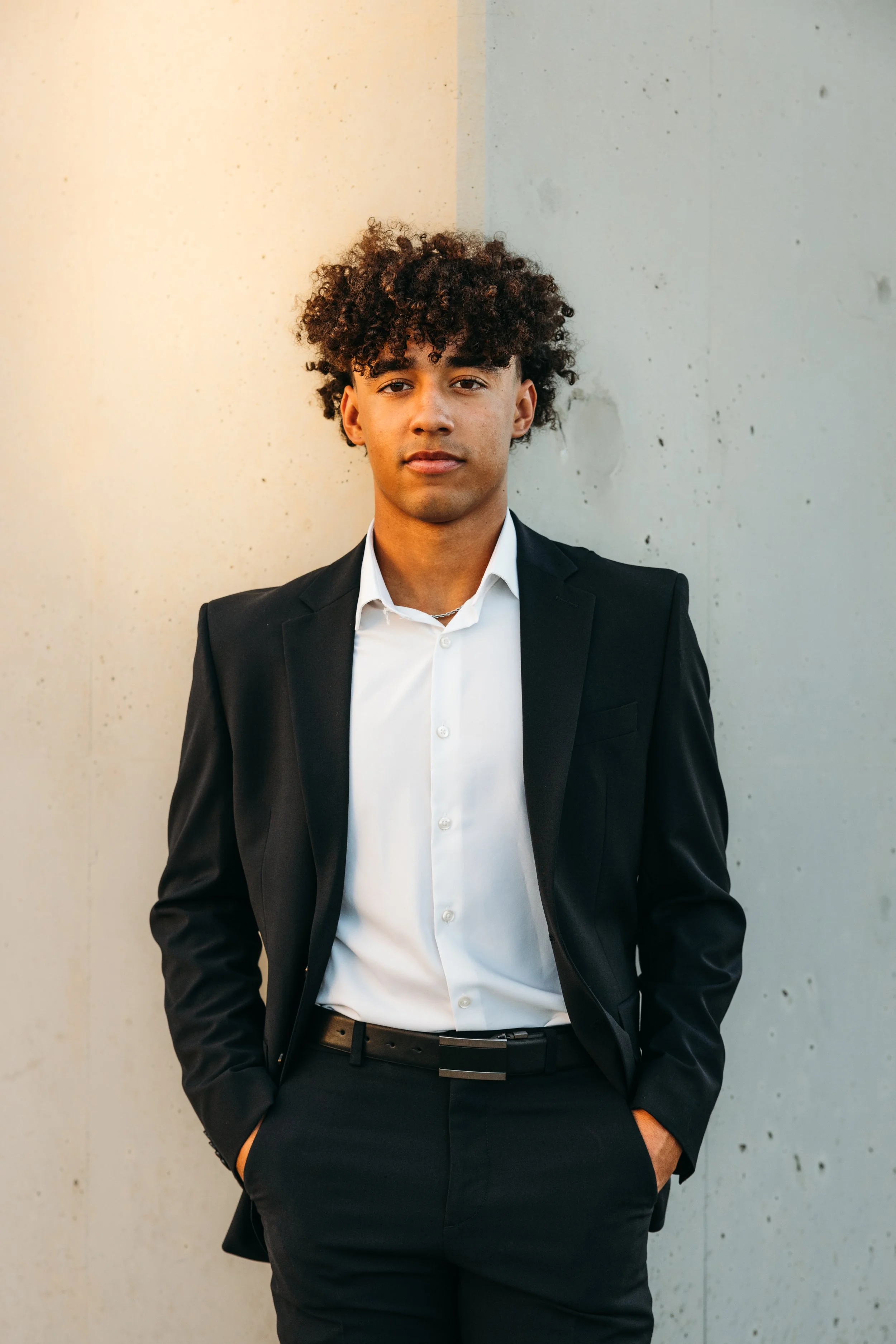 A young man with curly dark hair wearing a black suit, white shirt, and black belt, standing against a plain concrete wall.