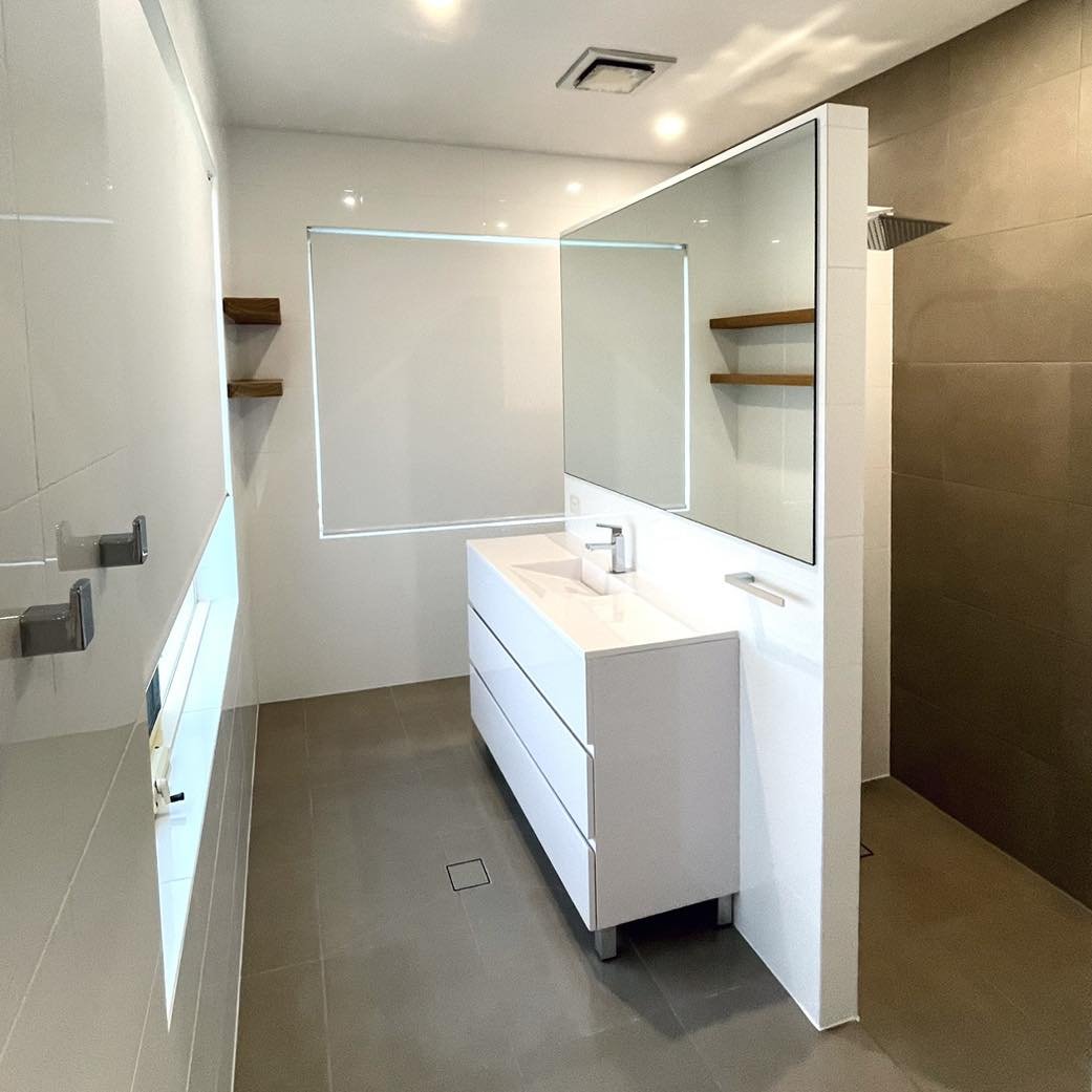 Modern ensuite bathroom renovation featuring a white wall-hung vanity, large mirror, floating timber shelves, walk-in shower, feature wall and floor tiles, a floating nib wall, and a concealed toilet.