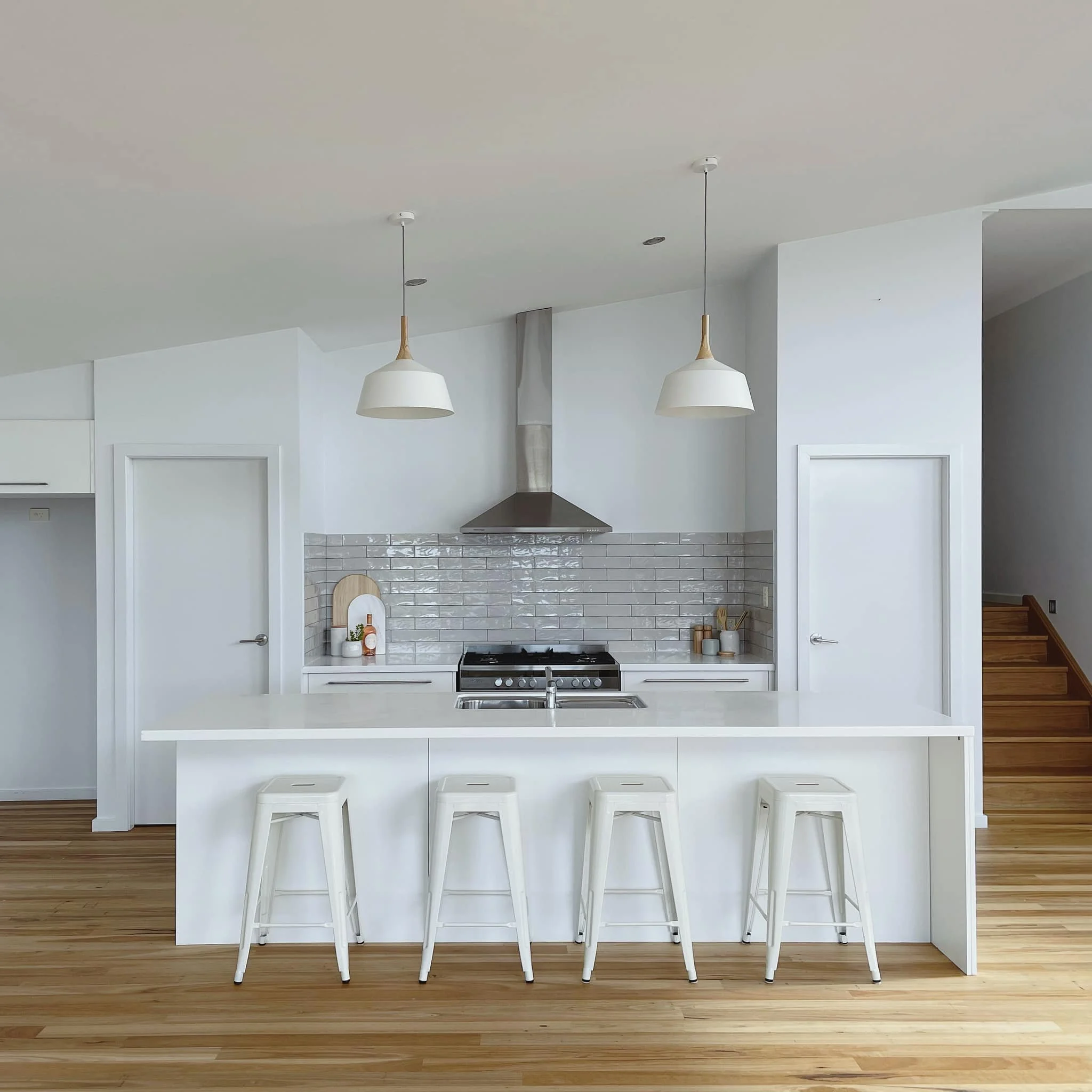 Modern white kitchen with timber floorboards, stainless steel stove, subway tile backsplash, hanging pendant lights, and a raked ceiling. Floor sanding completed and lighting updated as part of a minor renovation.